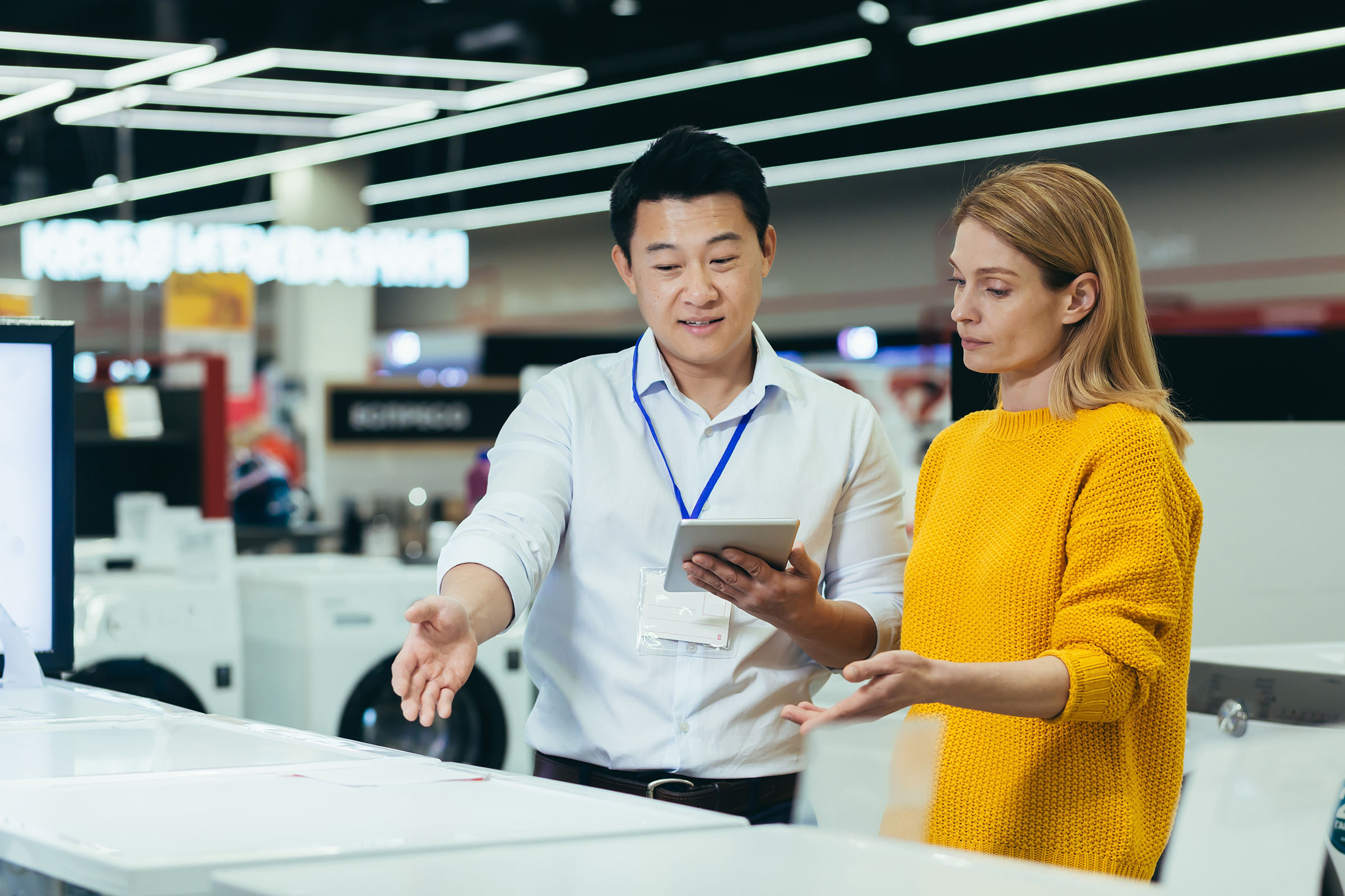 Sales associate showing a customer appliance options using a tablet in an electronics or home goods store.
