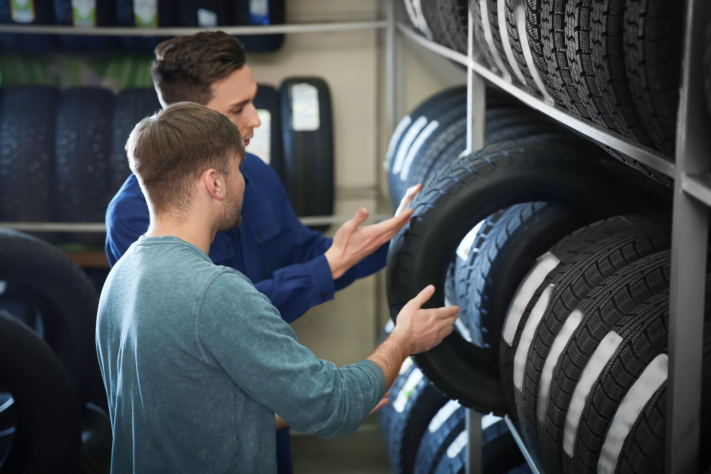 Two men examining car tires in a store, surrounded by shelves stocked with various tire options.