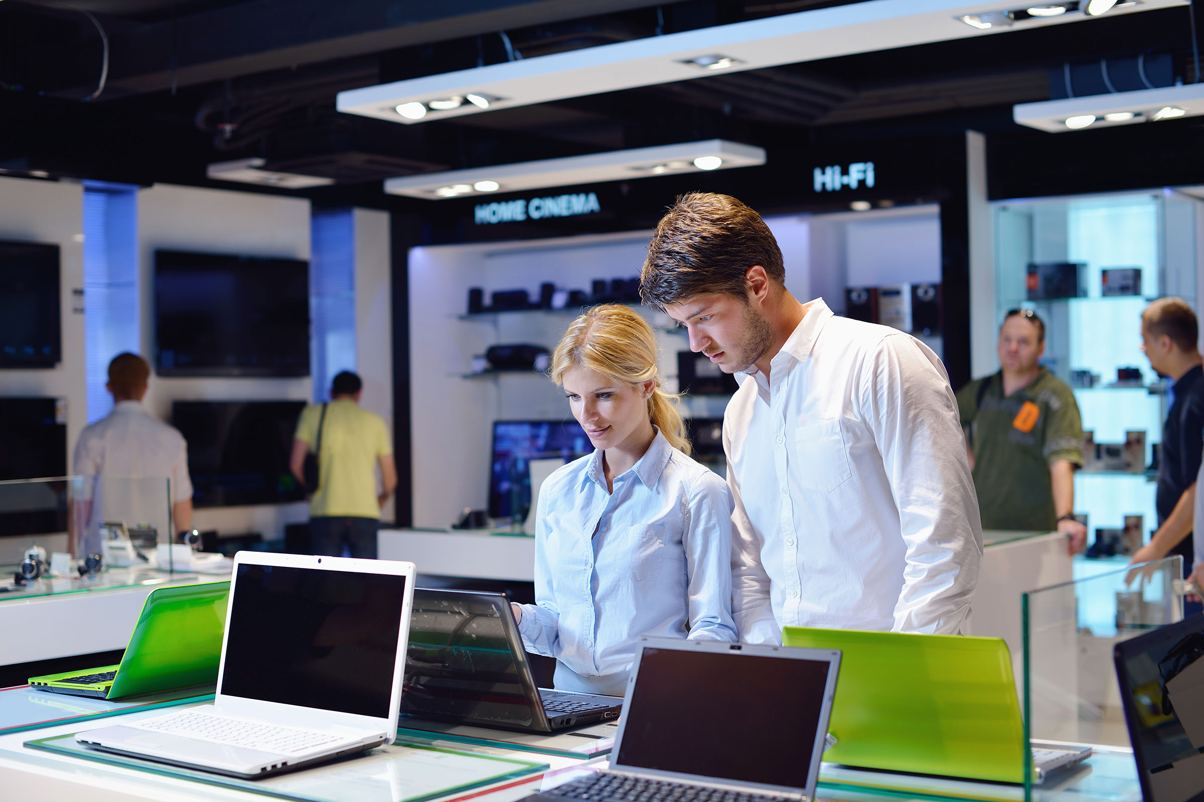 A man and a woman are looking at laptops in an electronics store with various gadgets and televisions displayed in the background.