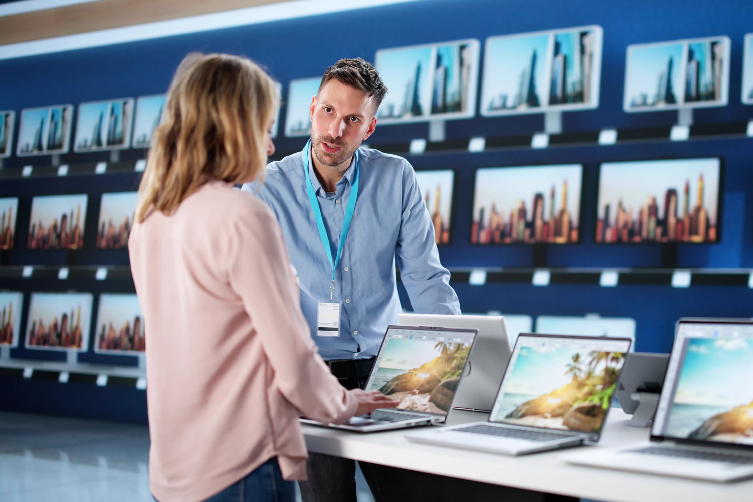 Woman speaking with a store employee while looking at laptops in an electronics store, with TV screens displayed on the wall behind them.