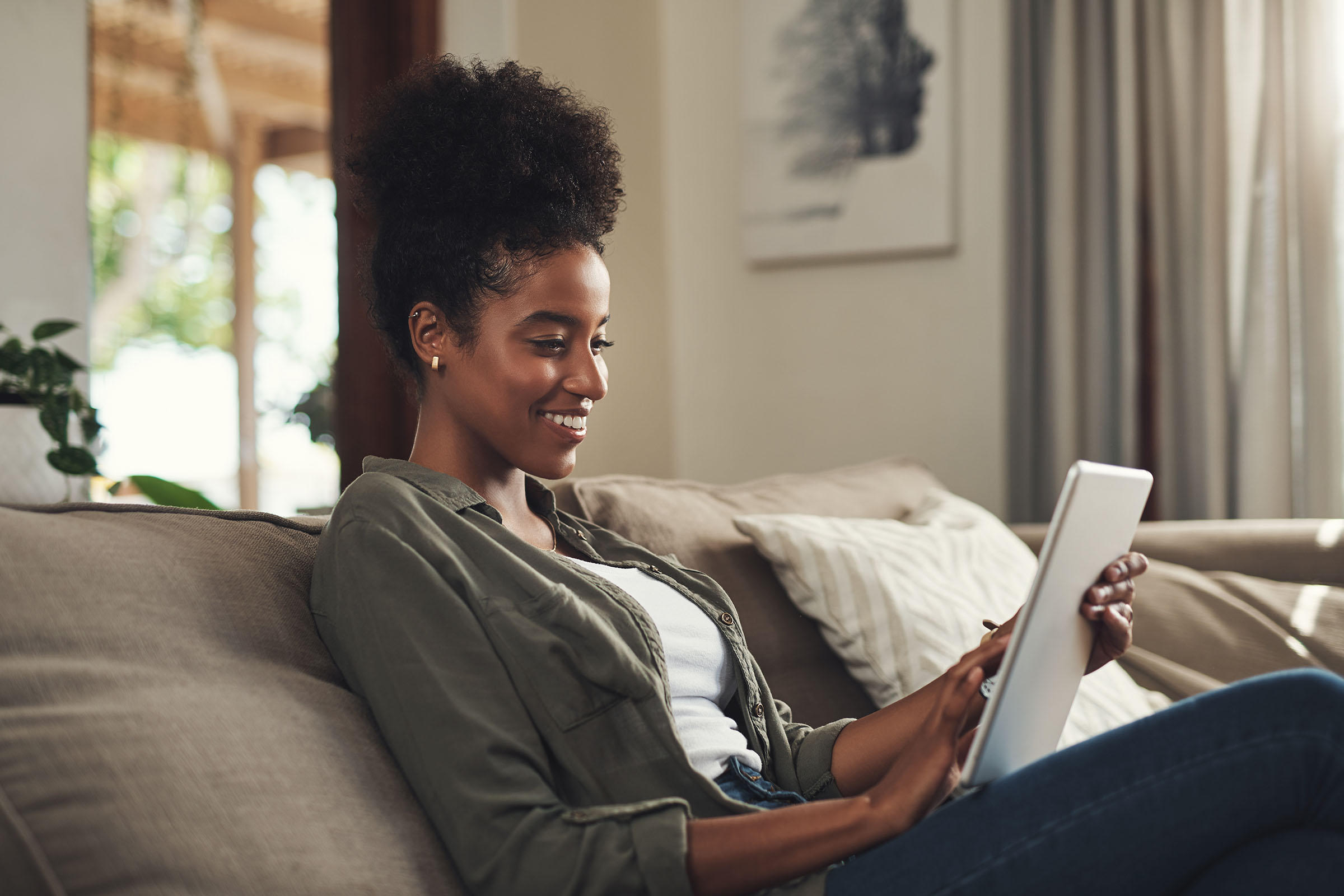 A woman sitting on a couch, smiling while using a tablet in a cozy living room.