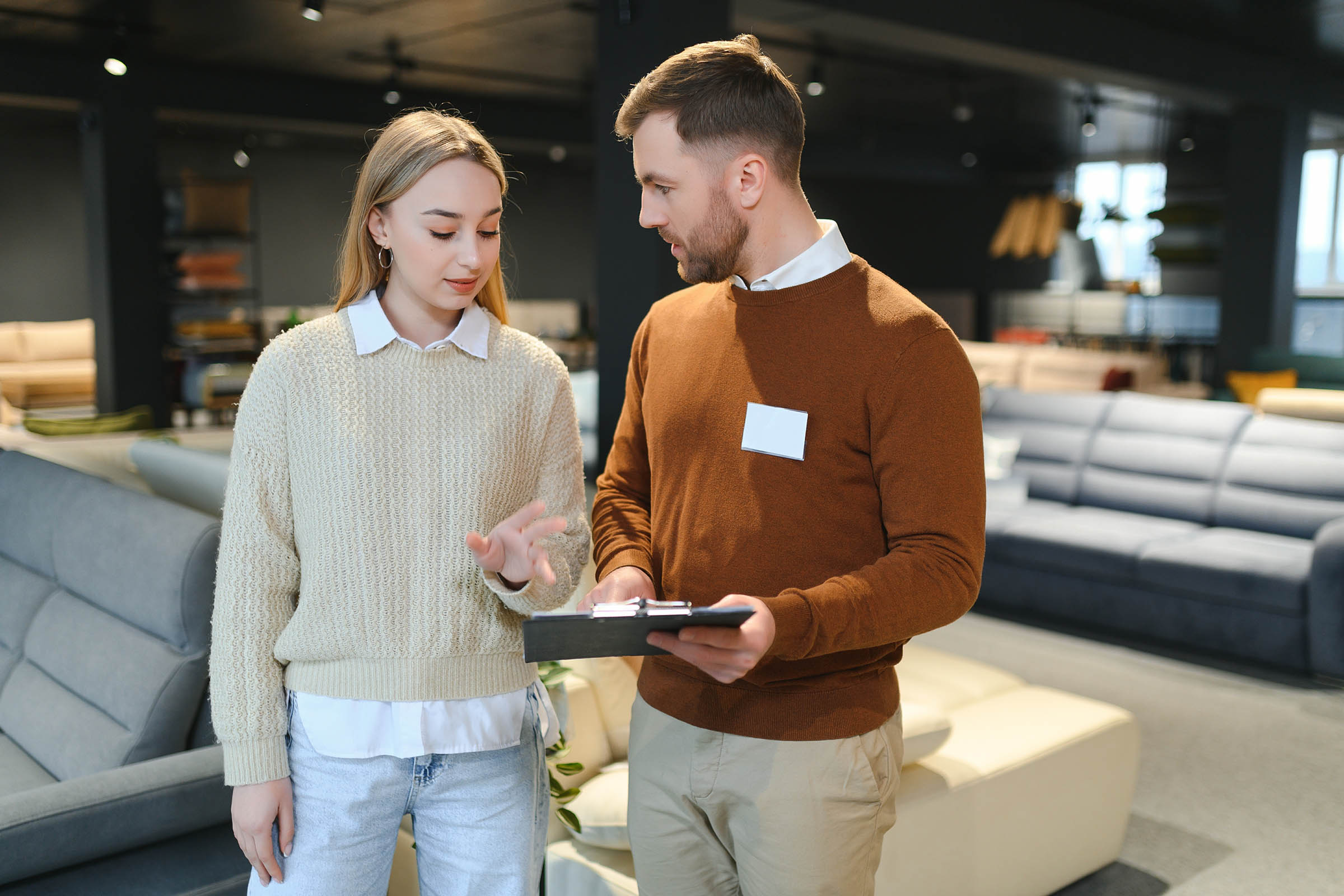 A woman and a man in a furniture store discuss while looking at a clipboard. Sofas are visible in the background.
