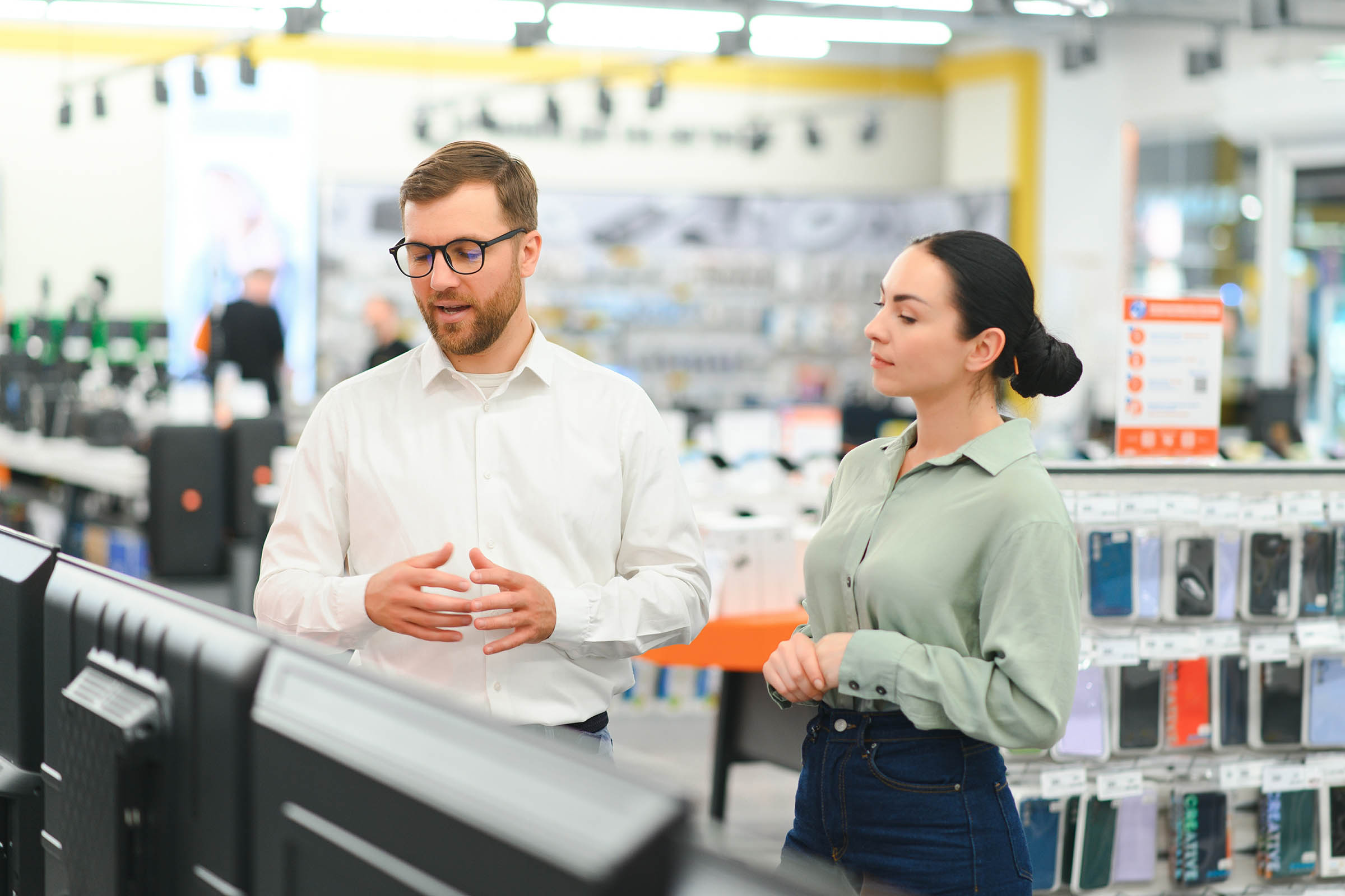 A man and woman discuss while standing in a bright electronics store, surrounded by various gadgets and products on display.