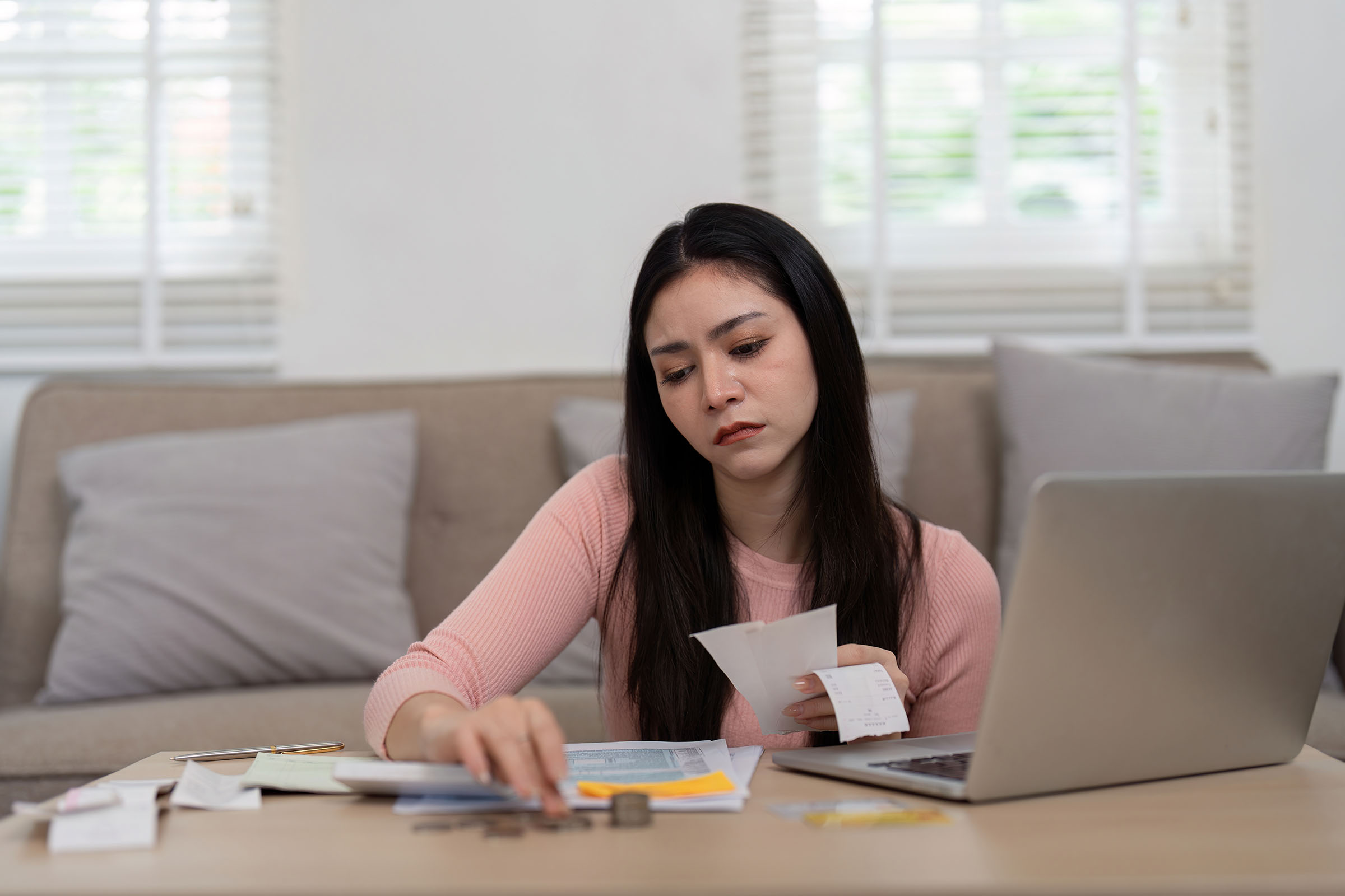 Woman sitting at a table, reviewing receipts and counting coins, with a laptop open, in front of a beige couch and white windows.
