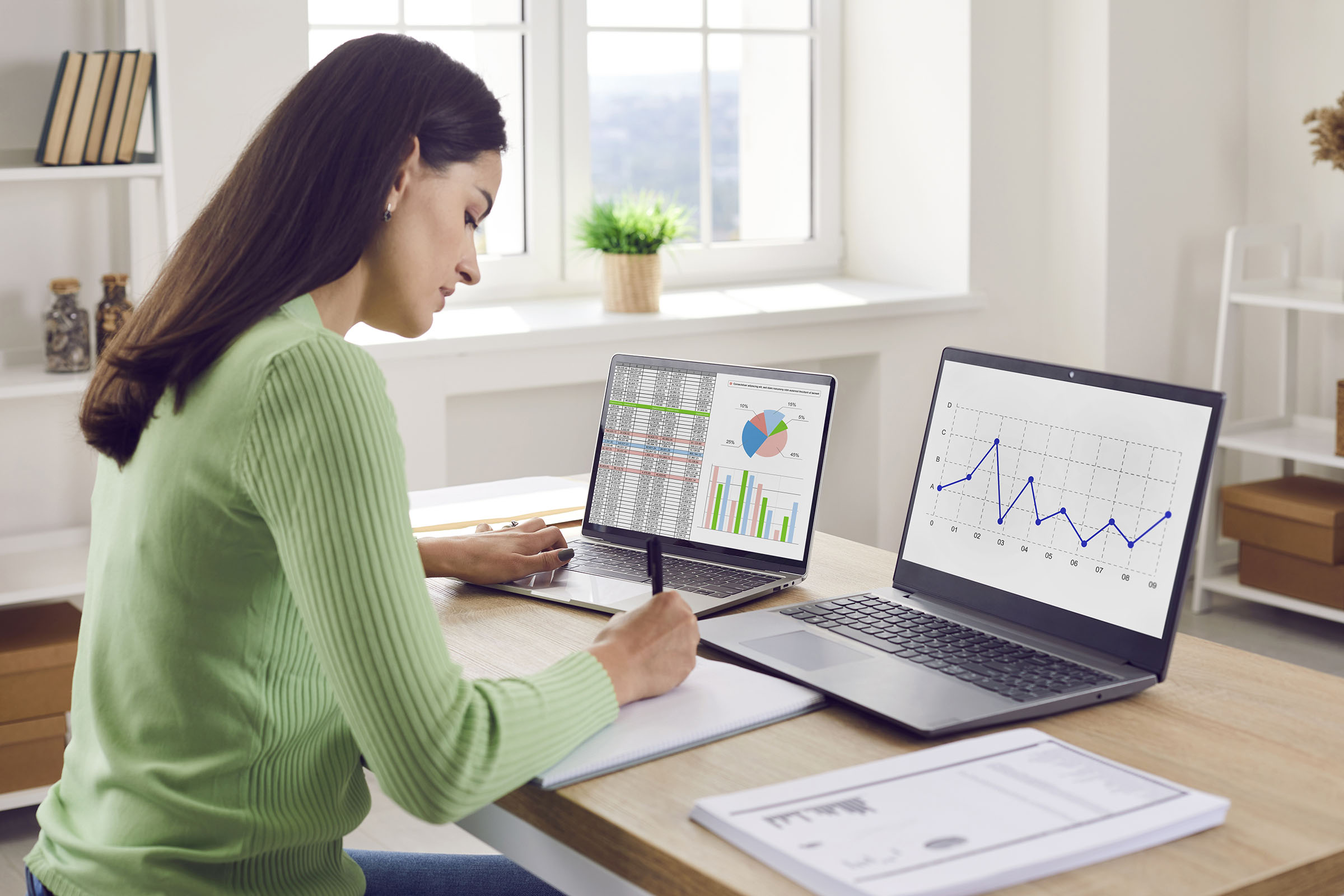 Woman in green sweater working at a desk with two laptops displaying charts and graphs, writing on a notepad.