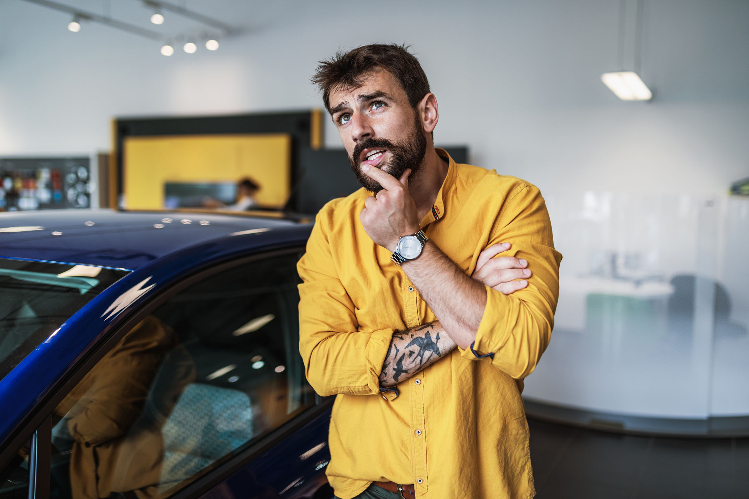 A man in a yellow shirt with a thoughtful expression stands next to a blue car in a brightly lit showroom.