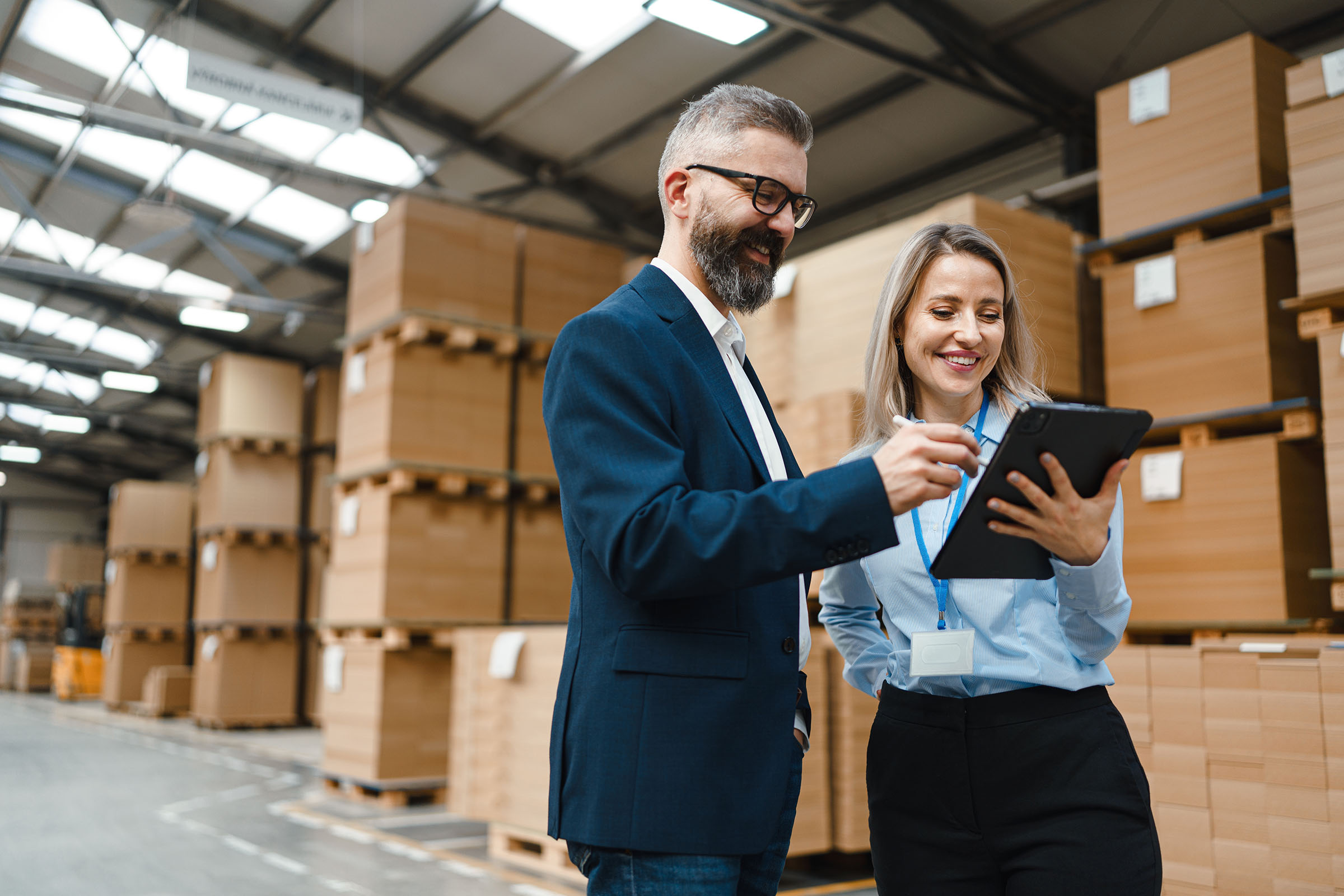 A man and a woman smiling as they review information on a tablet in a warehouse with stacked boxes.