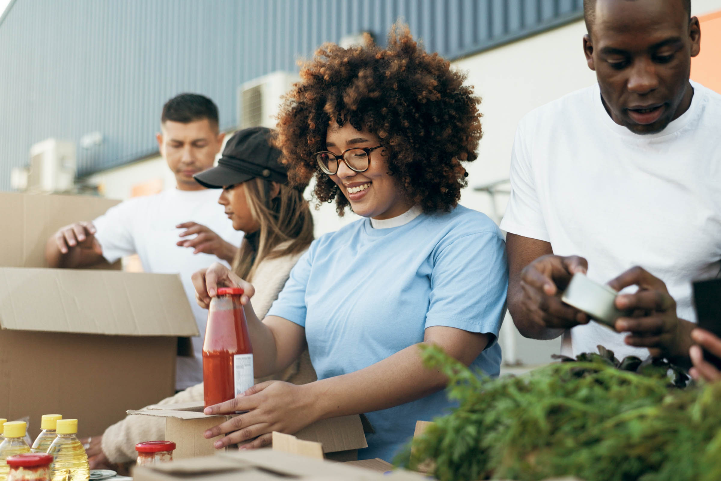 A group of volunteers smiling and packing food items, including jars and canned goods, into boxes outdoors.