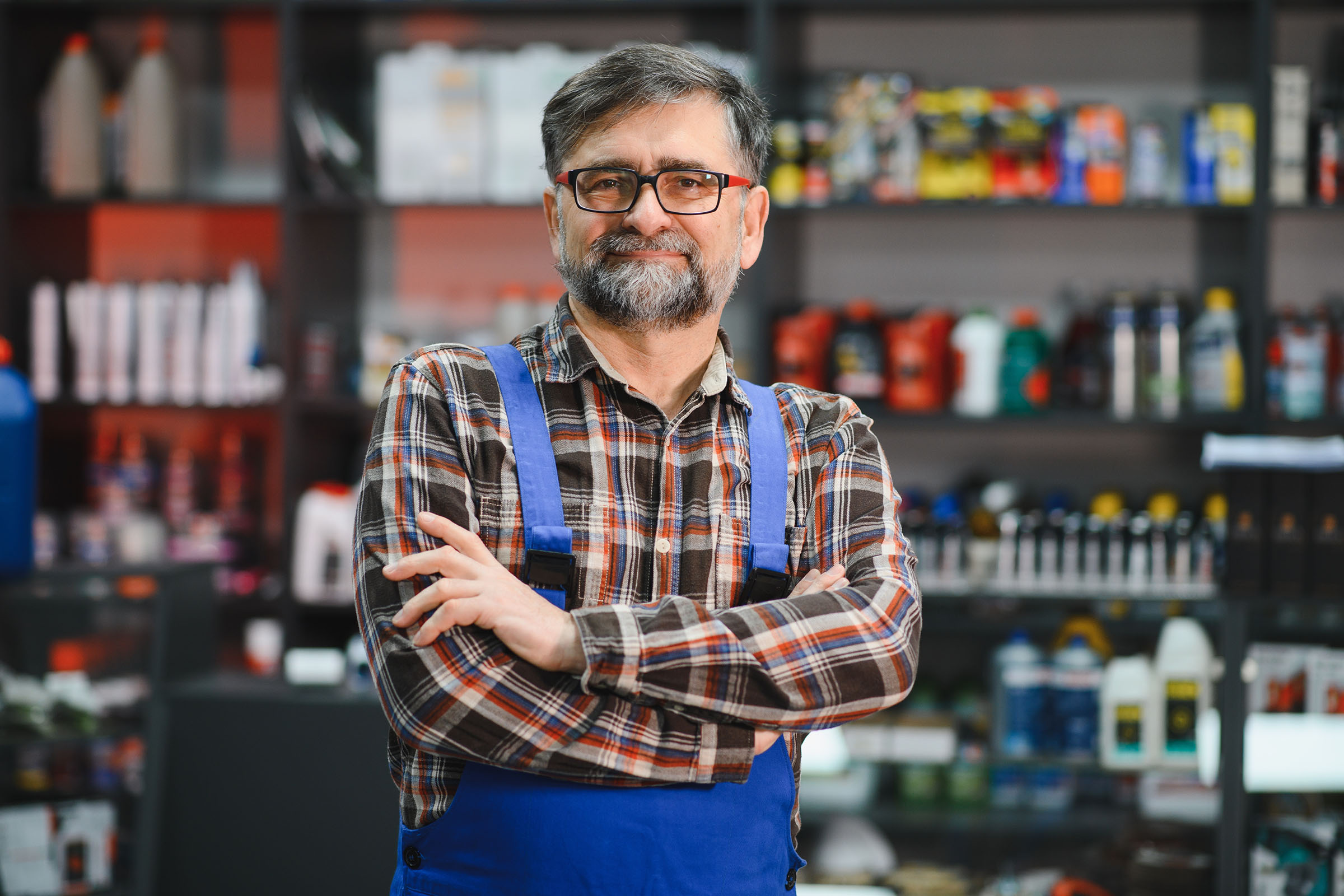 Man in plaid shirt and blue overalls stands with arms crossed in a store with shelves of automotive products in the background.