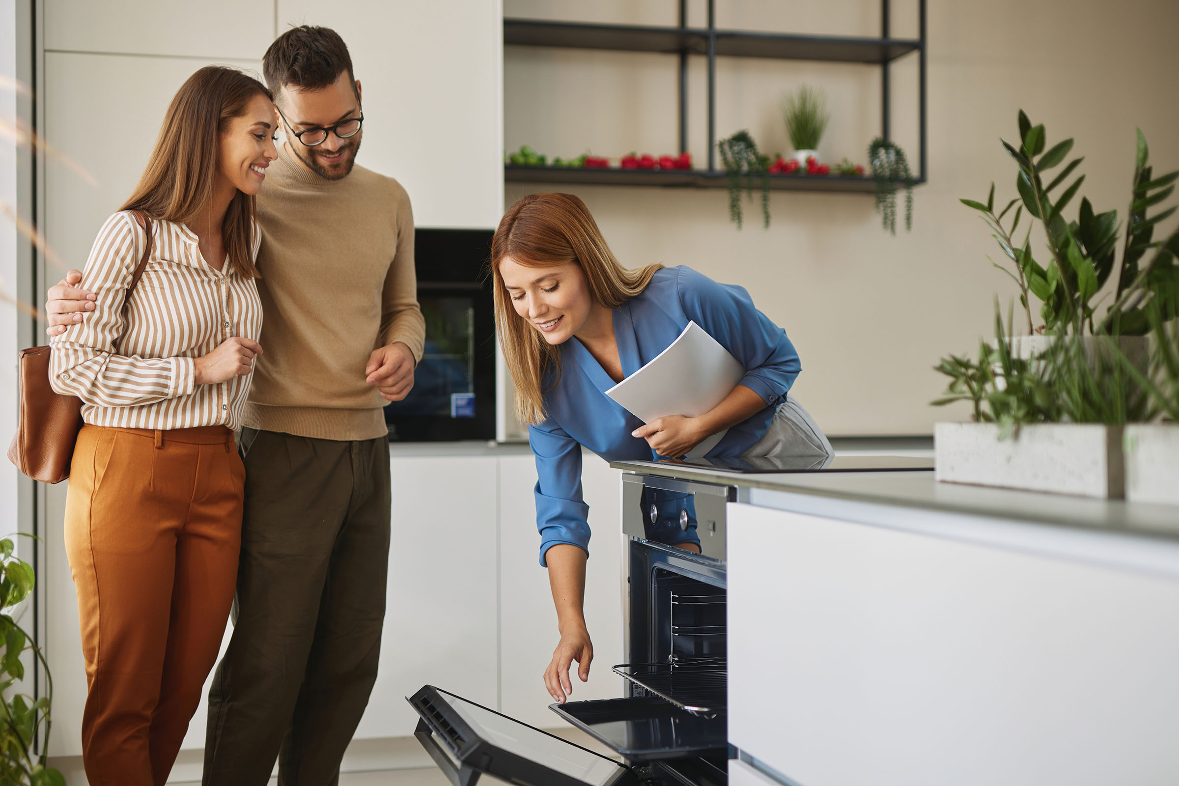 Saleswoman showing an open oven to a smiling couple in a modern kitchen showroom with plants and contemporary decor.