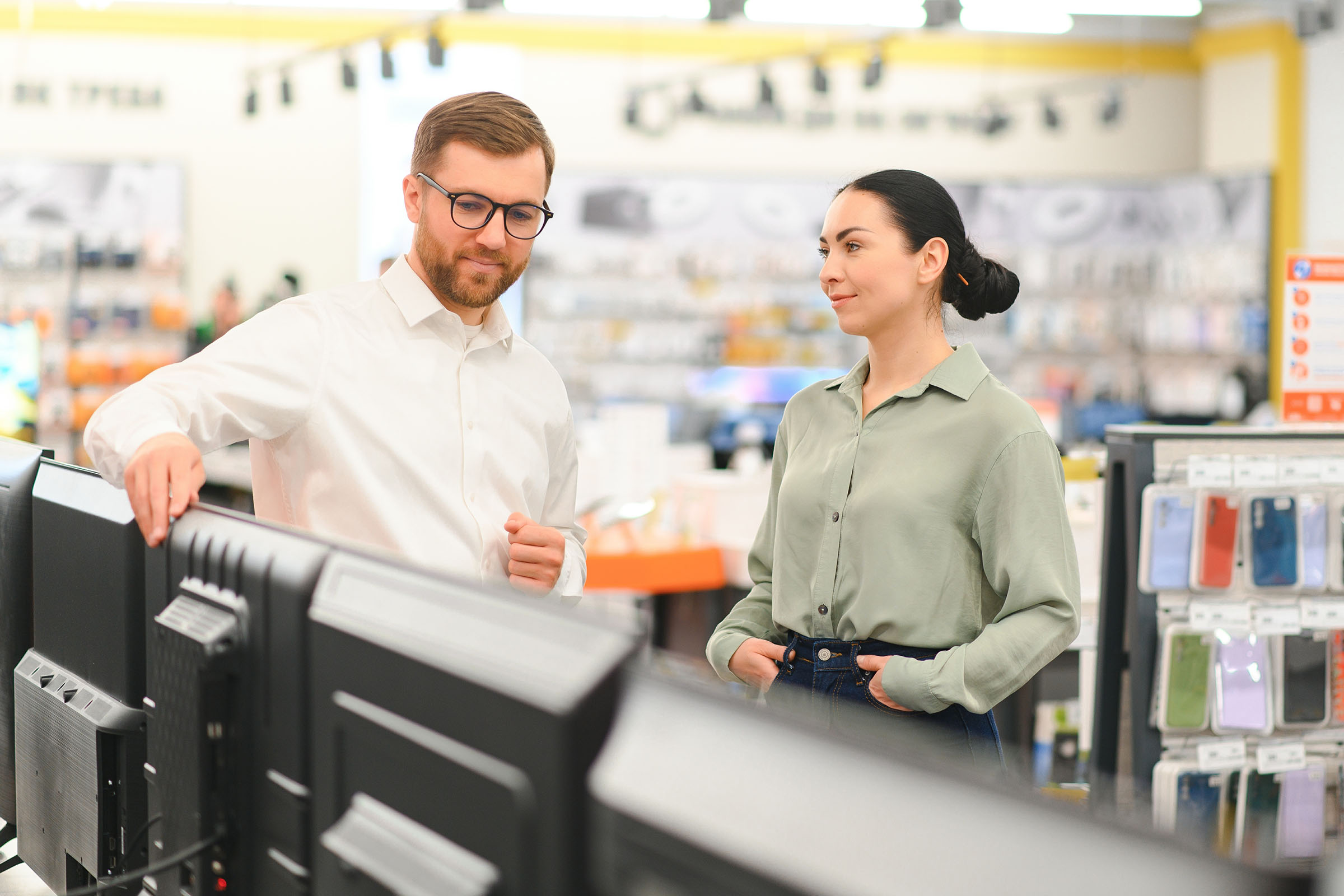 A man in a white shirt and glasses explains something to a woman in a green blouse in an electronics store, standing near monitors.