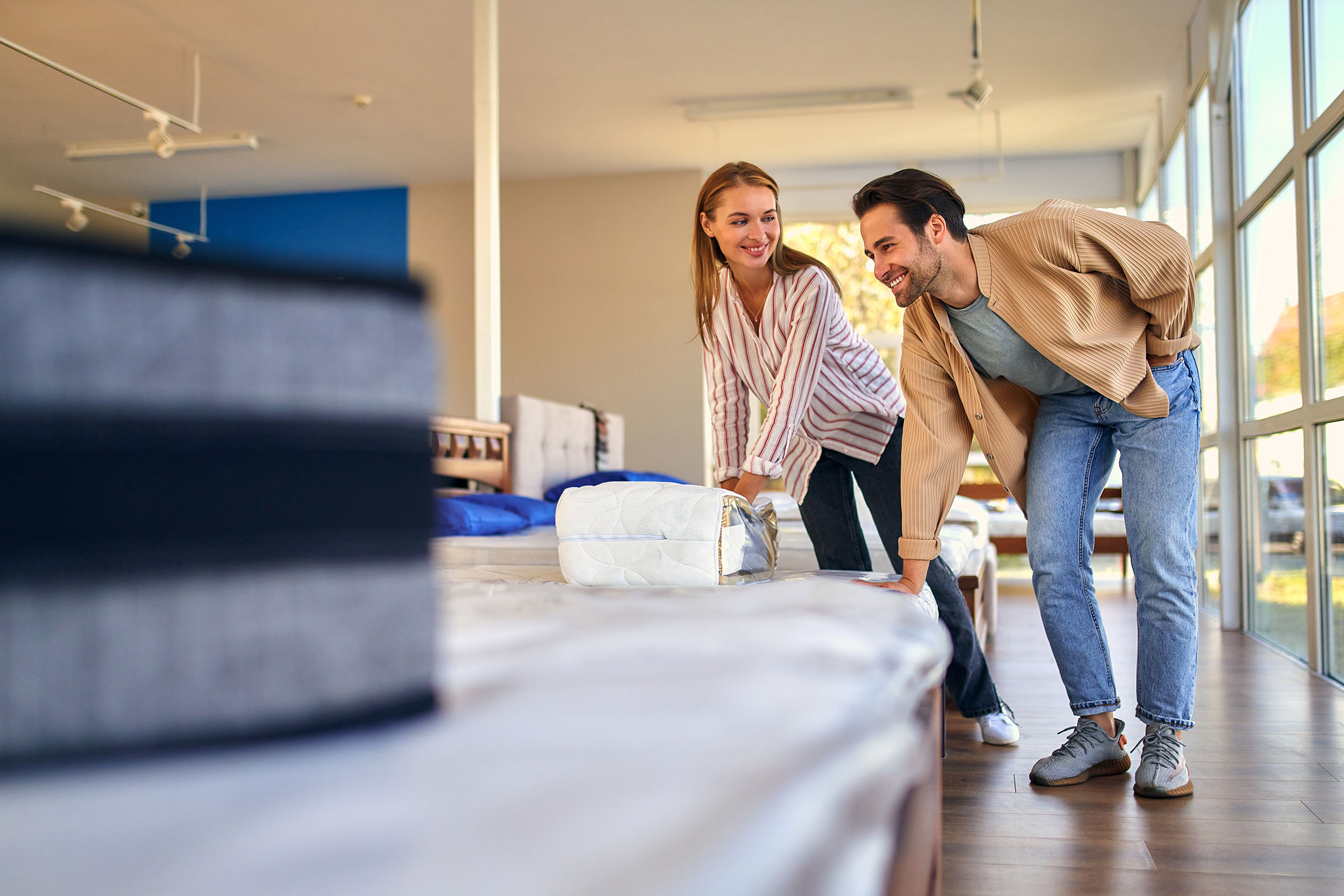 Smiling couple testing mattresses by pressing on them in a brightly lit showroom with large windows.