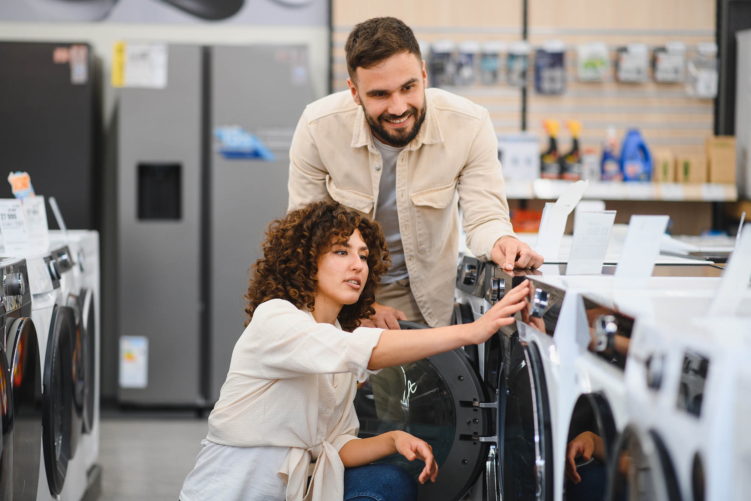 A couple examines washing machines in an appliance store, with the woman adjusting controls and the man overseeing.