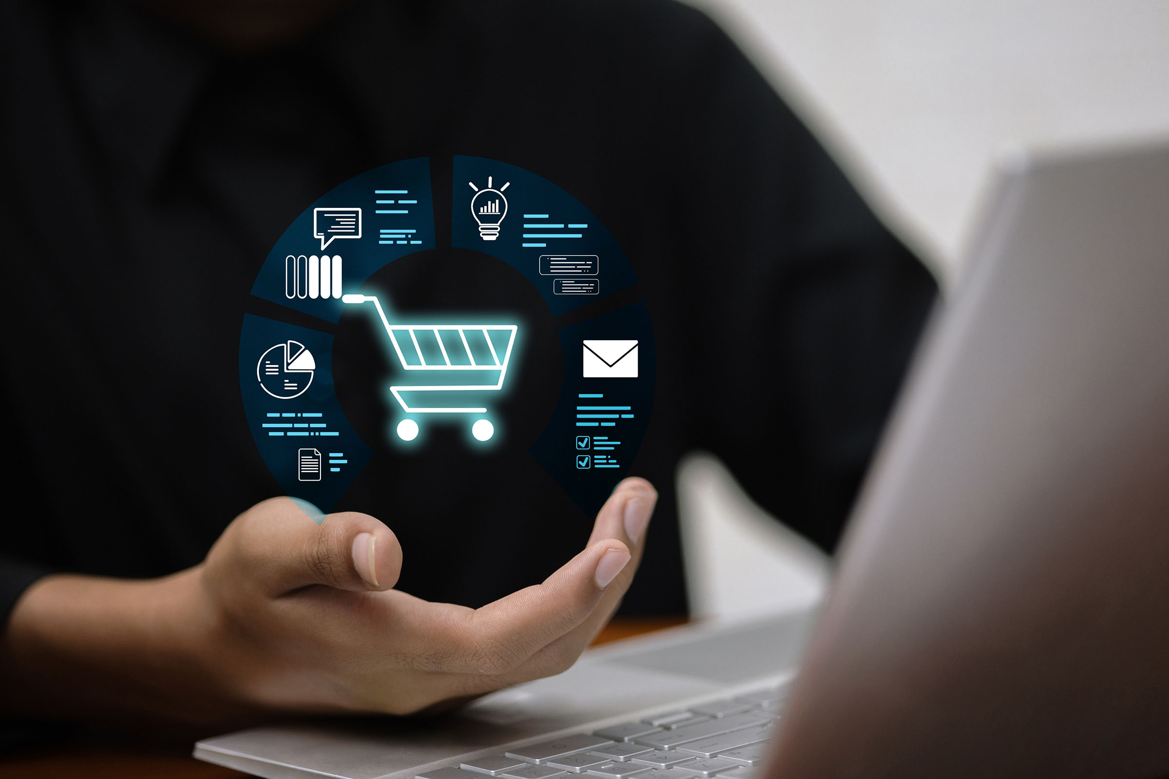 A person holds a digital shopping cart icon surrounded by analytics and communication symbols, in front of a laptop.