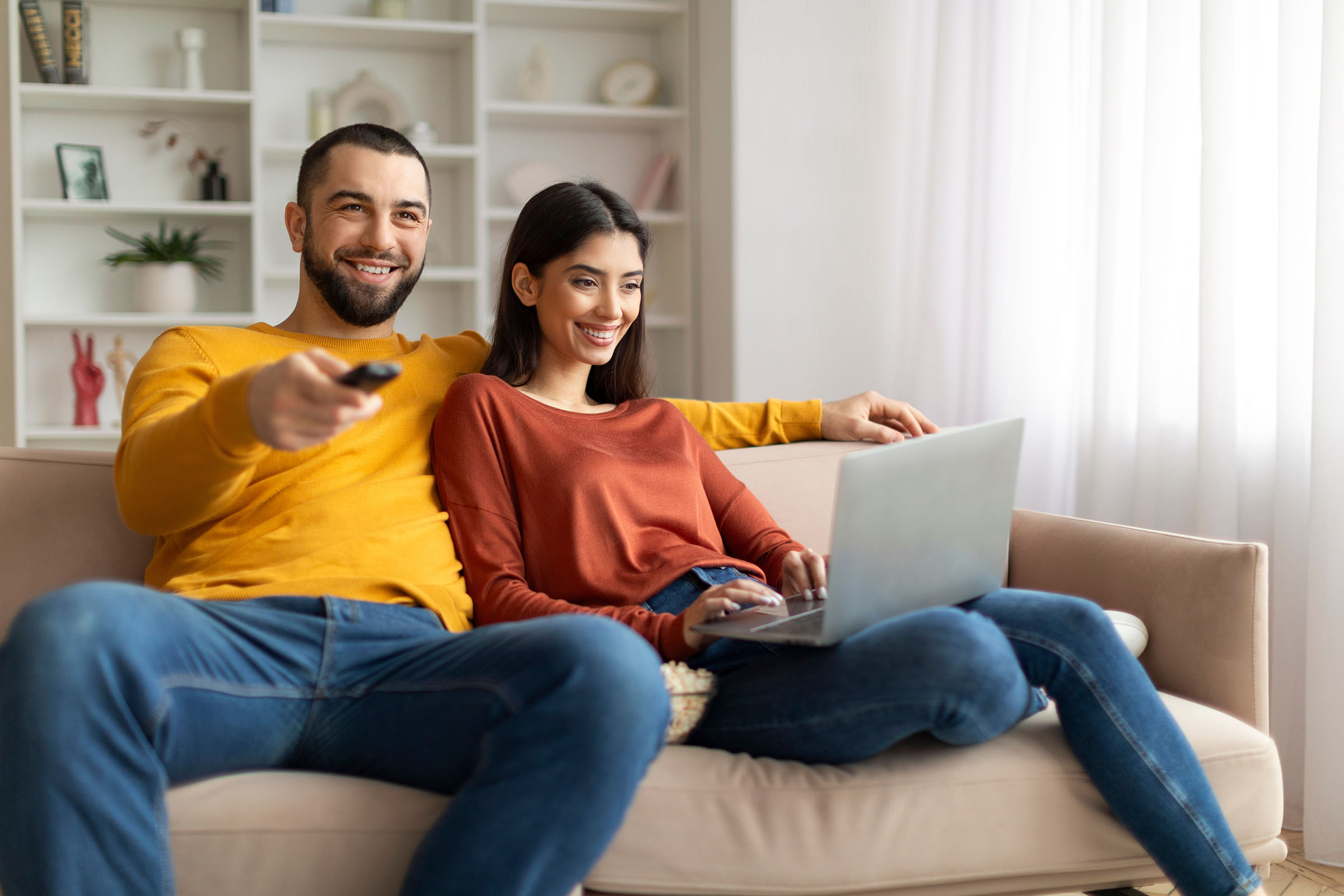 Smiling couple sitting on a sofa, with one using a laptop and the other holding a TV remote, appearing relaxed in a cozy living room.
