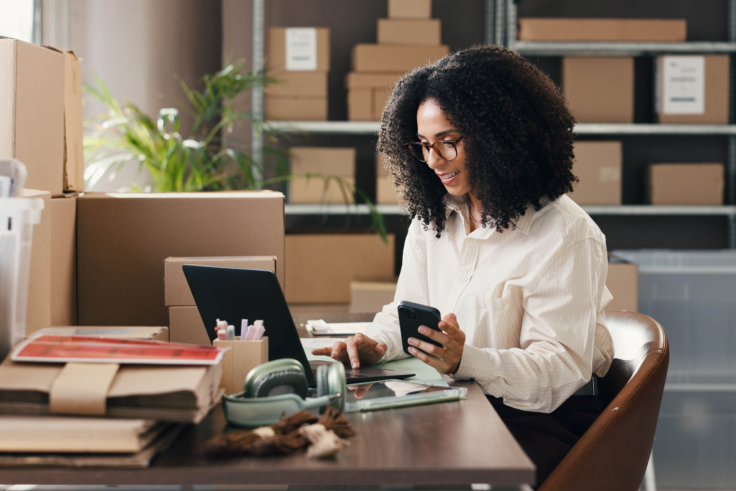 A woman with curly hair and glasses works at a desk with a laptop, surrounded by cardboard boxes in a warehouse office setting.