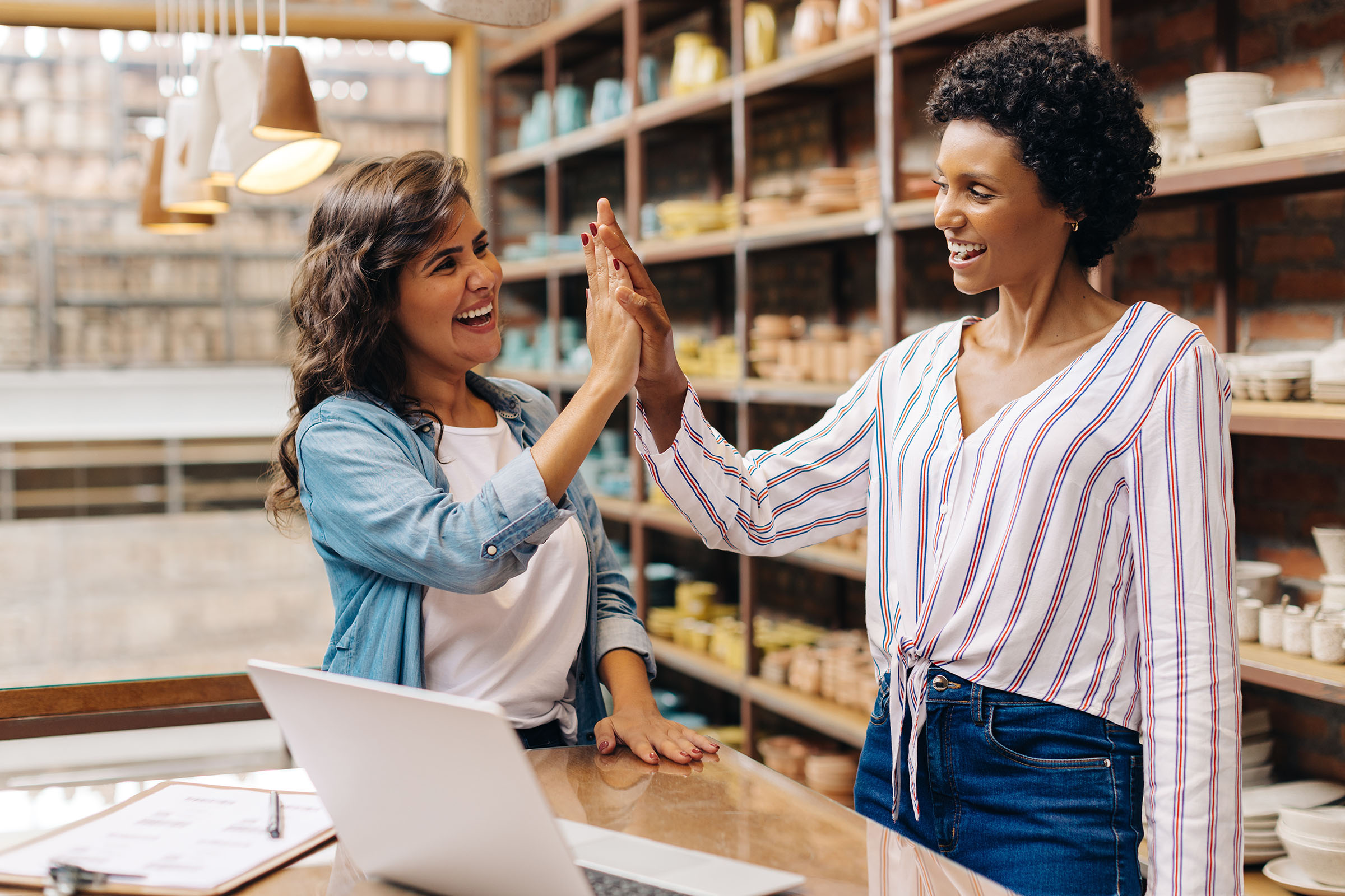 Two women smiling and high-fiving in a pottery store, standing at a counter with a laptop and clipboard. Shelves of pottery are in the background.