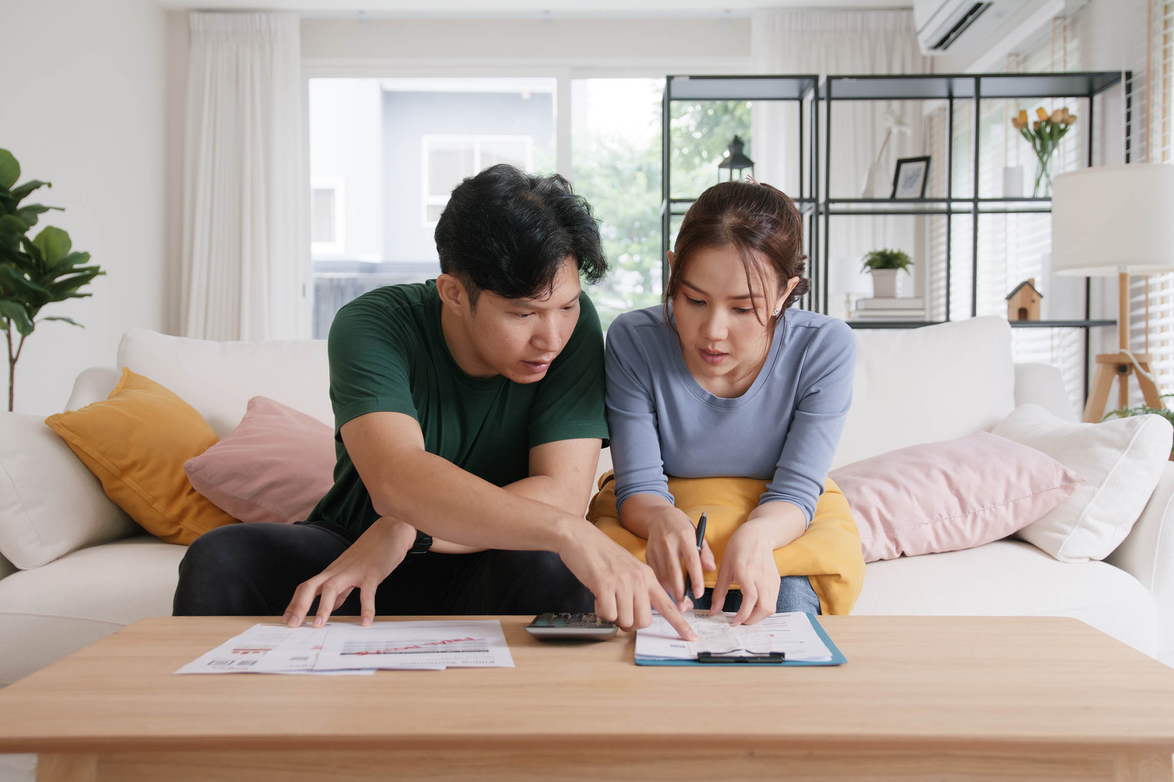 A couple sitting on a couch, discussing documents on a table, with a calculator nearby in a bright living room.