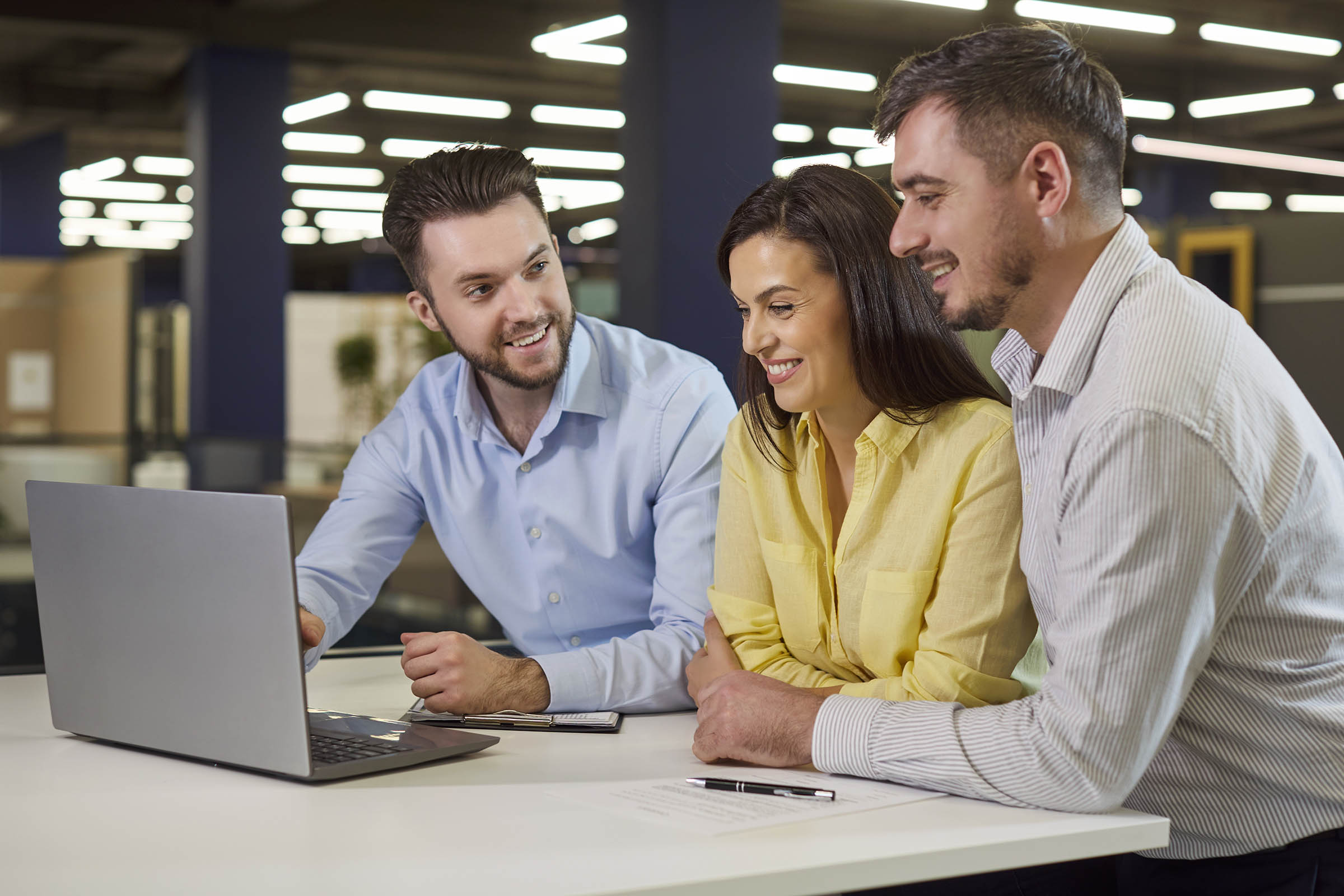 Three colleagues smiling and discussing work at a desk, looking at a laptop in a modern office environment.