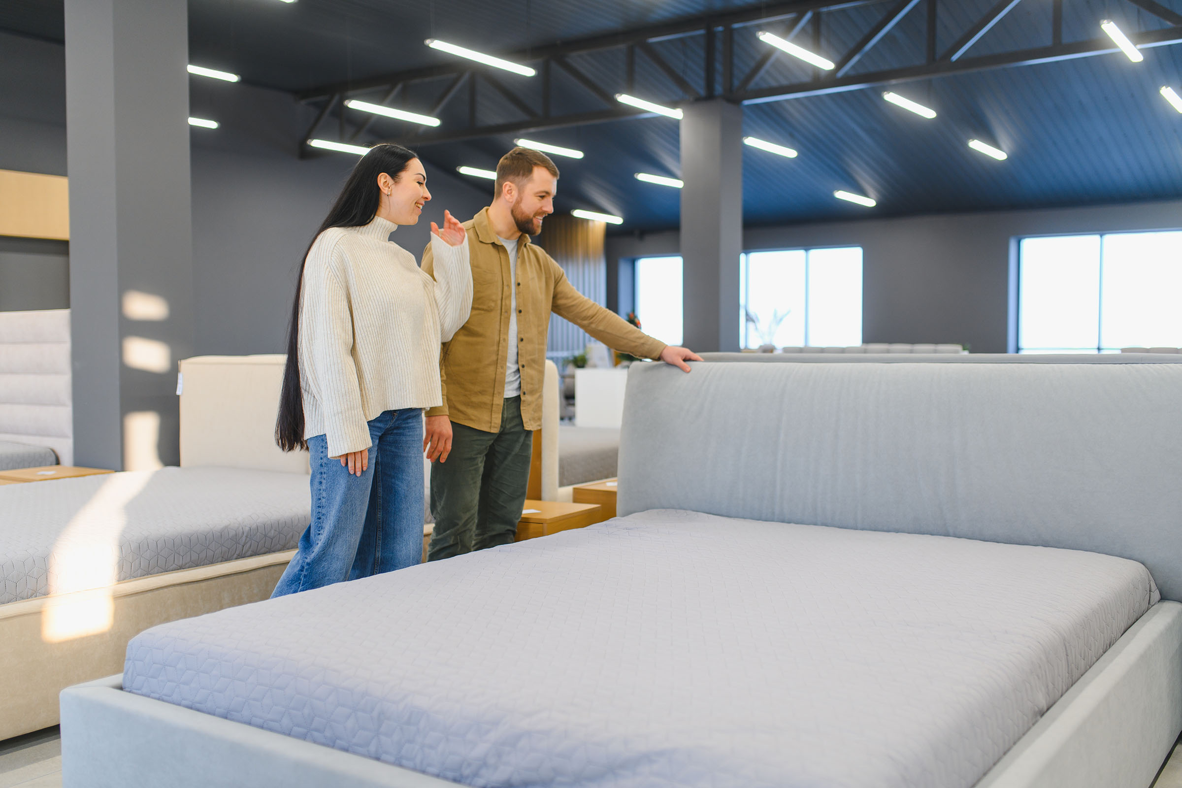A couple in a furniture store looks at a large bed with a smile. The store has modern lighting and several other beds on display.