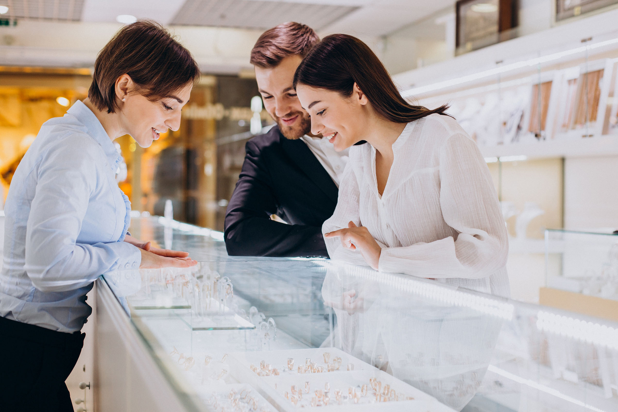 Smiling couple looking at rings in a jewelry store display case with assistance from a saleswoman.