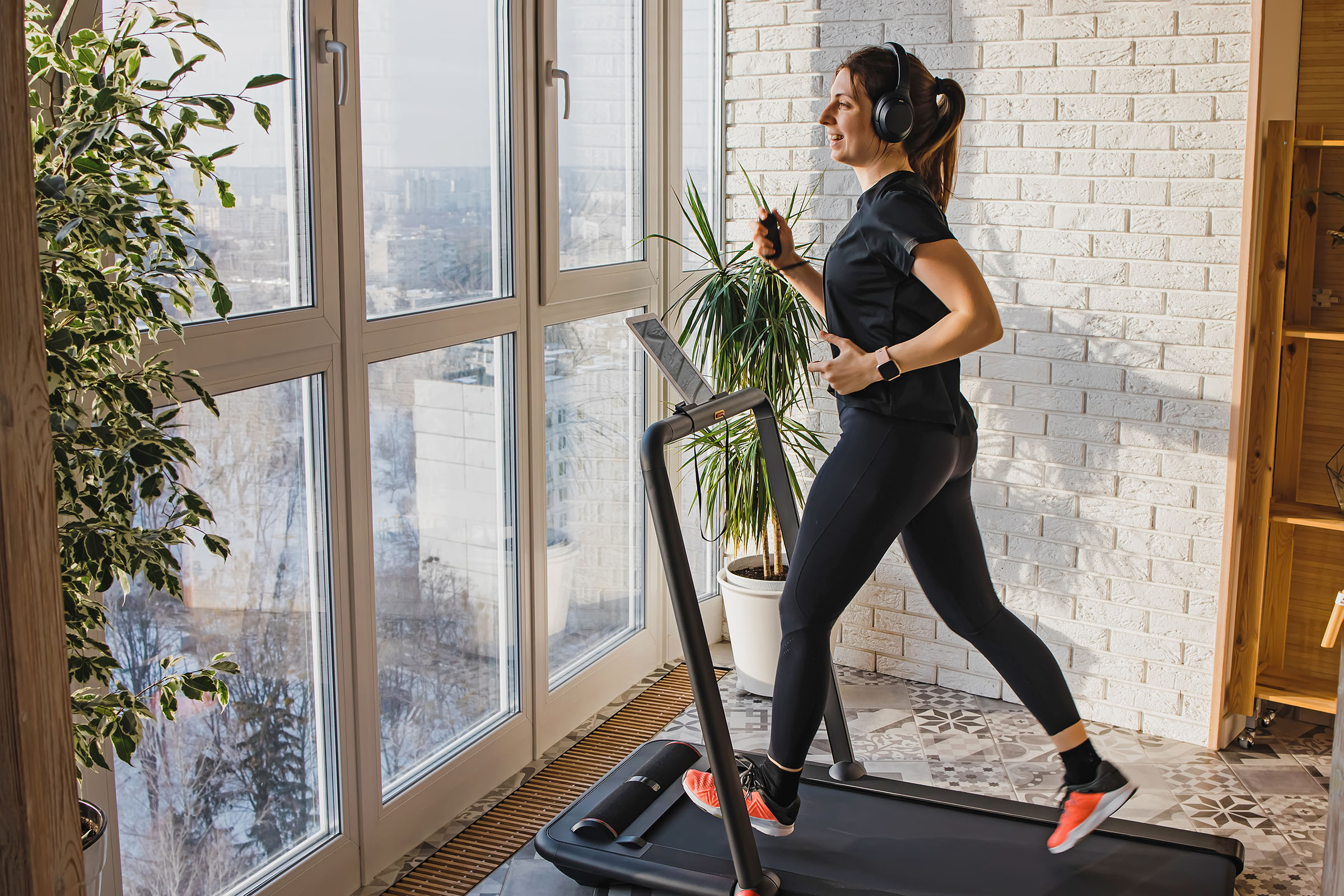 A woman runs on a treadmill at home near a large window, wearing headphones and athletic clothing, with a green plant in the background.