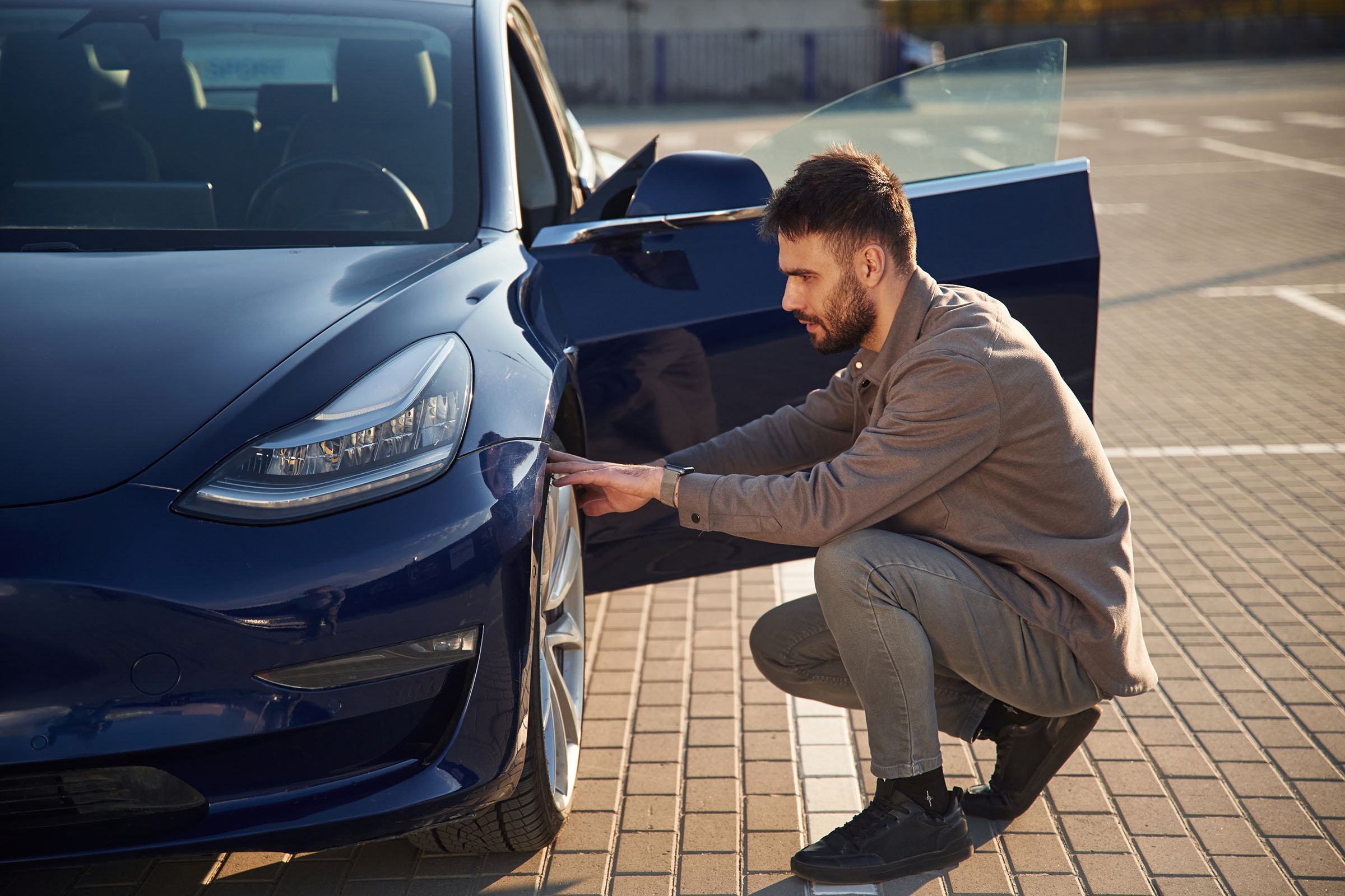 Man crouching to inspect the front bumper of a blue car in a parking lot with the driver's door open.