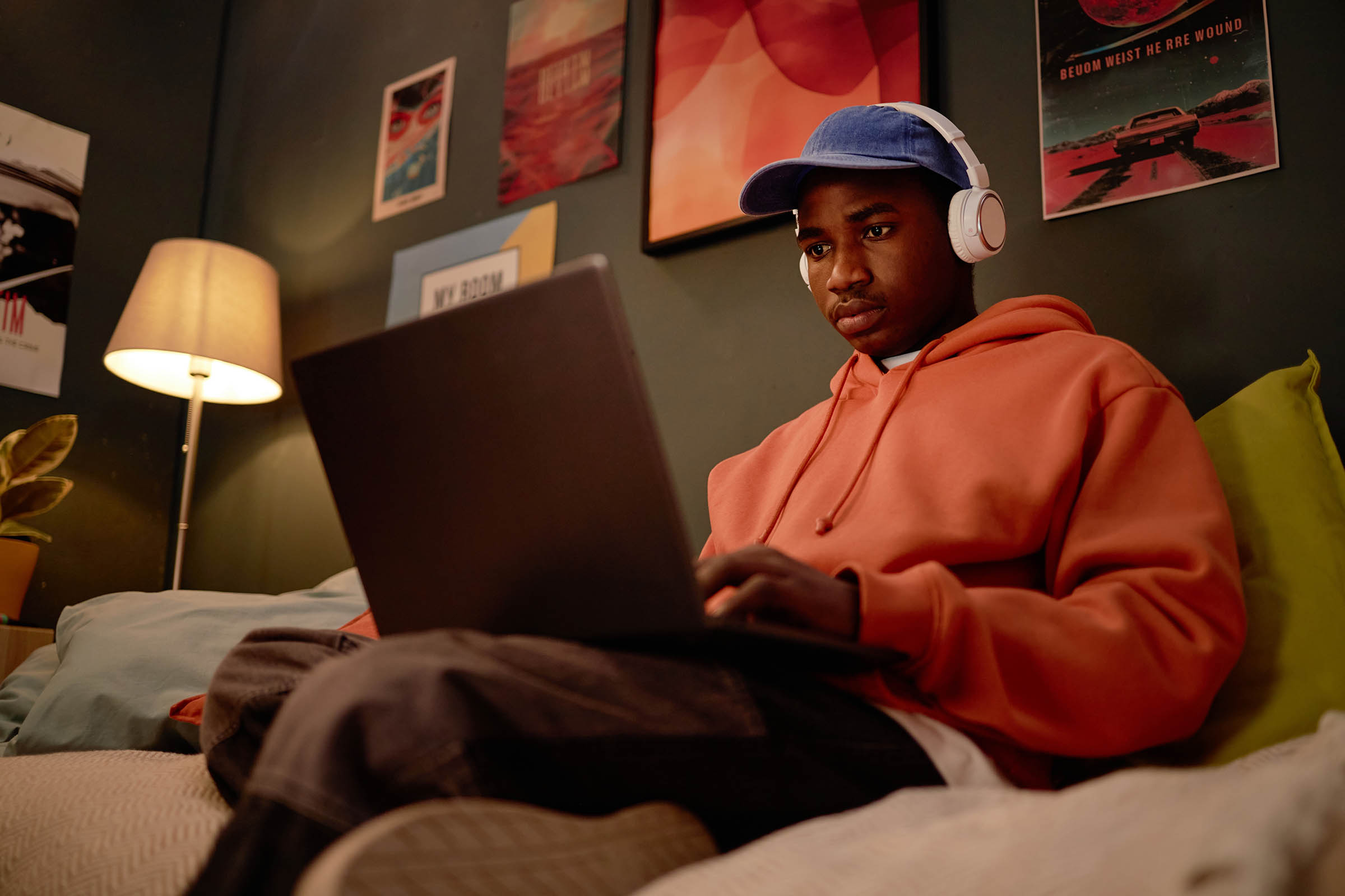 Person in a hoodie and cap sitting on a bed, using a laptop with headphones on, surrounded by wall posters and a lamp.