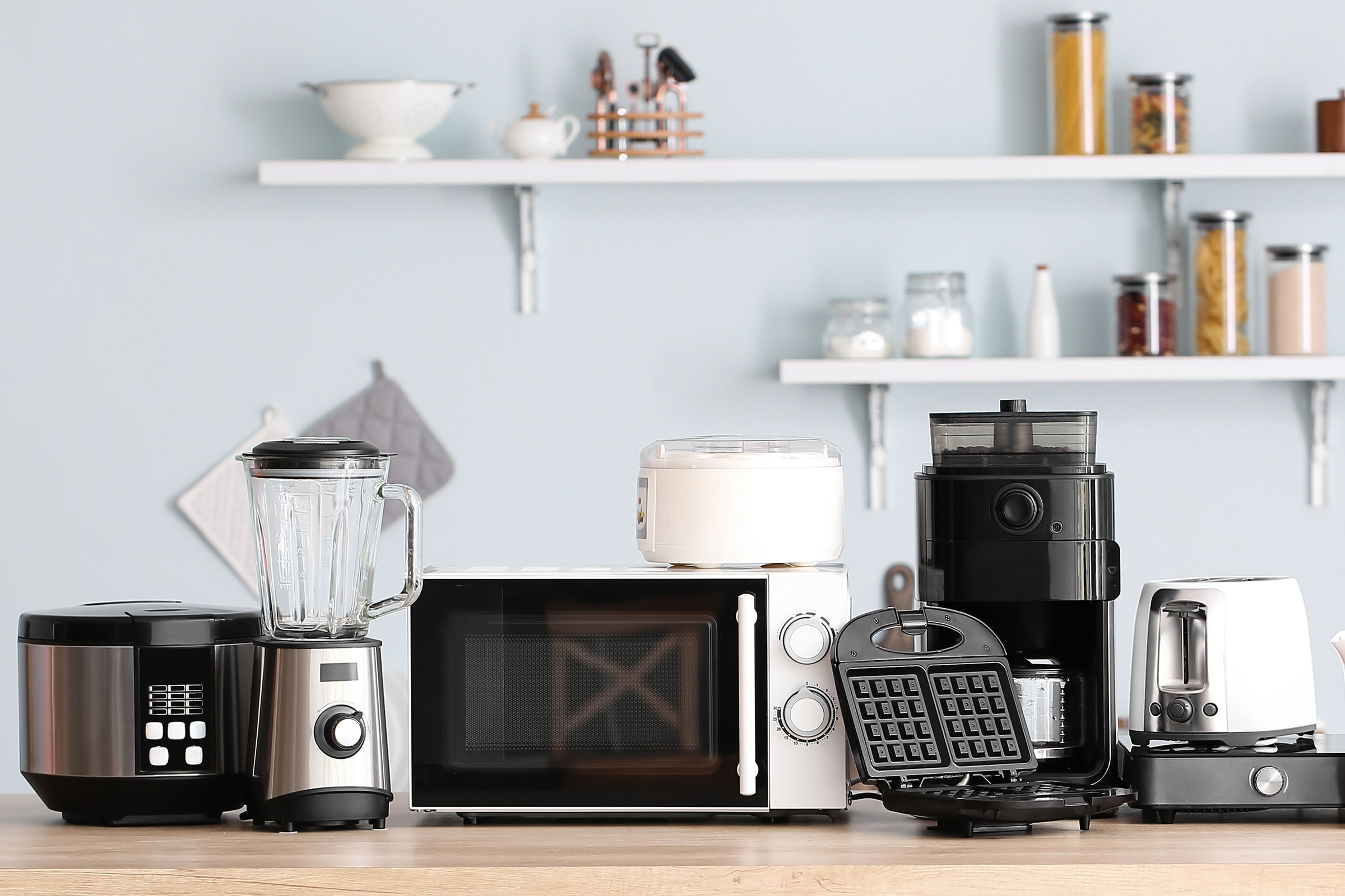 Variety of kitchen appliances on a counter, including a rice cooker, blender, microwave, ice cream maker, coffee maker, waffle maker, toaster, and hot plate.