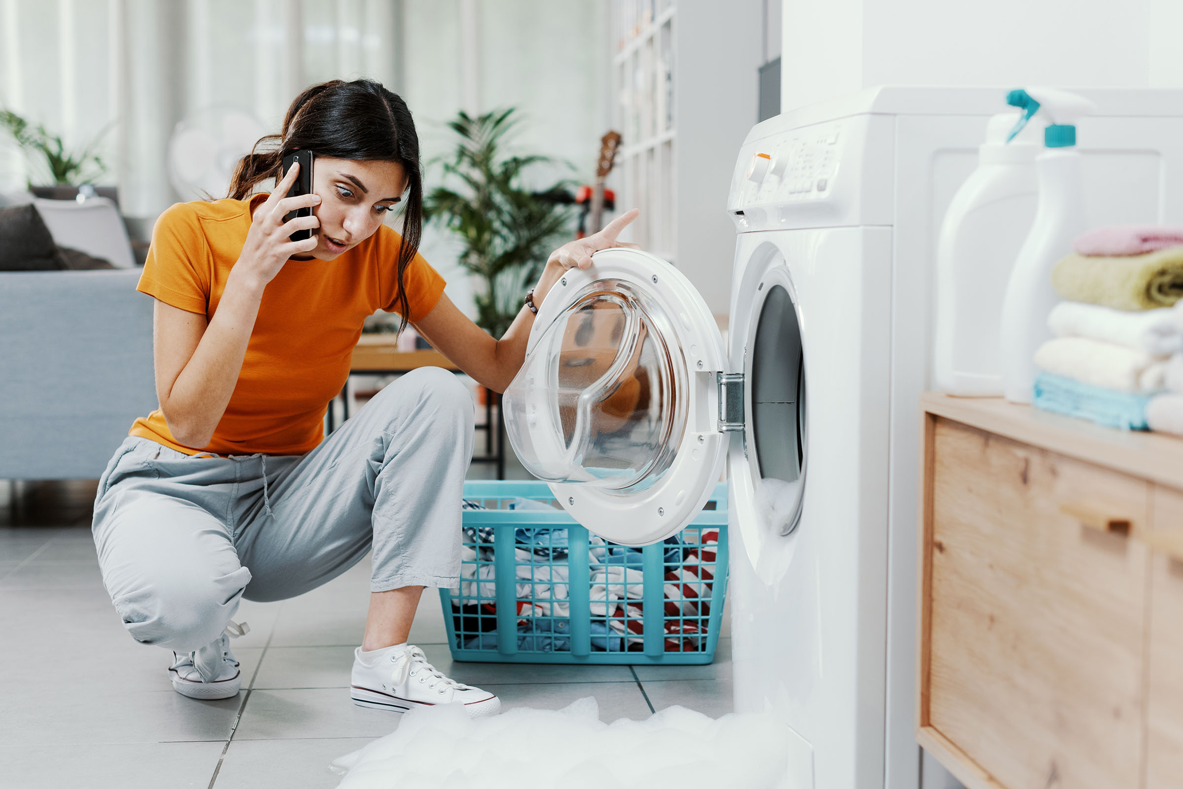 Concerned woman on the phone kneeling next to a washing machine overflowing with soap suds onto the floor.