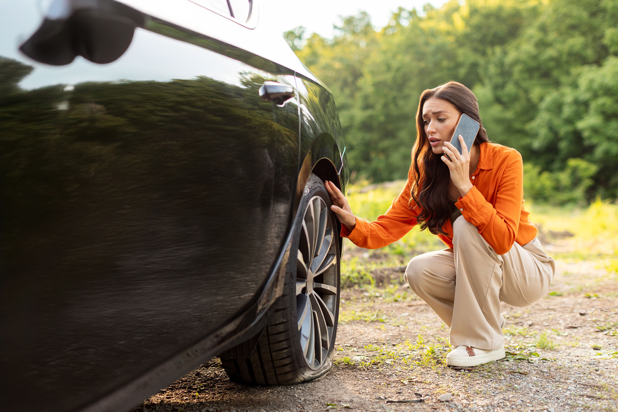 Concerned woman on the phone examining a flat tire on a black car parked on a dirt road near a wooded area.