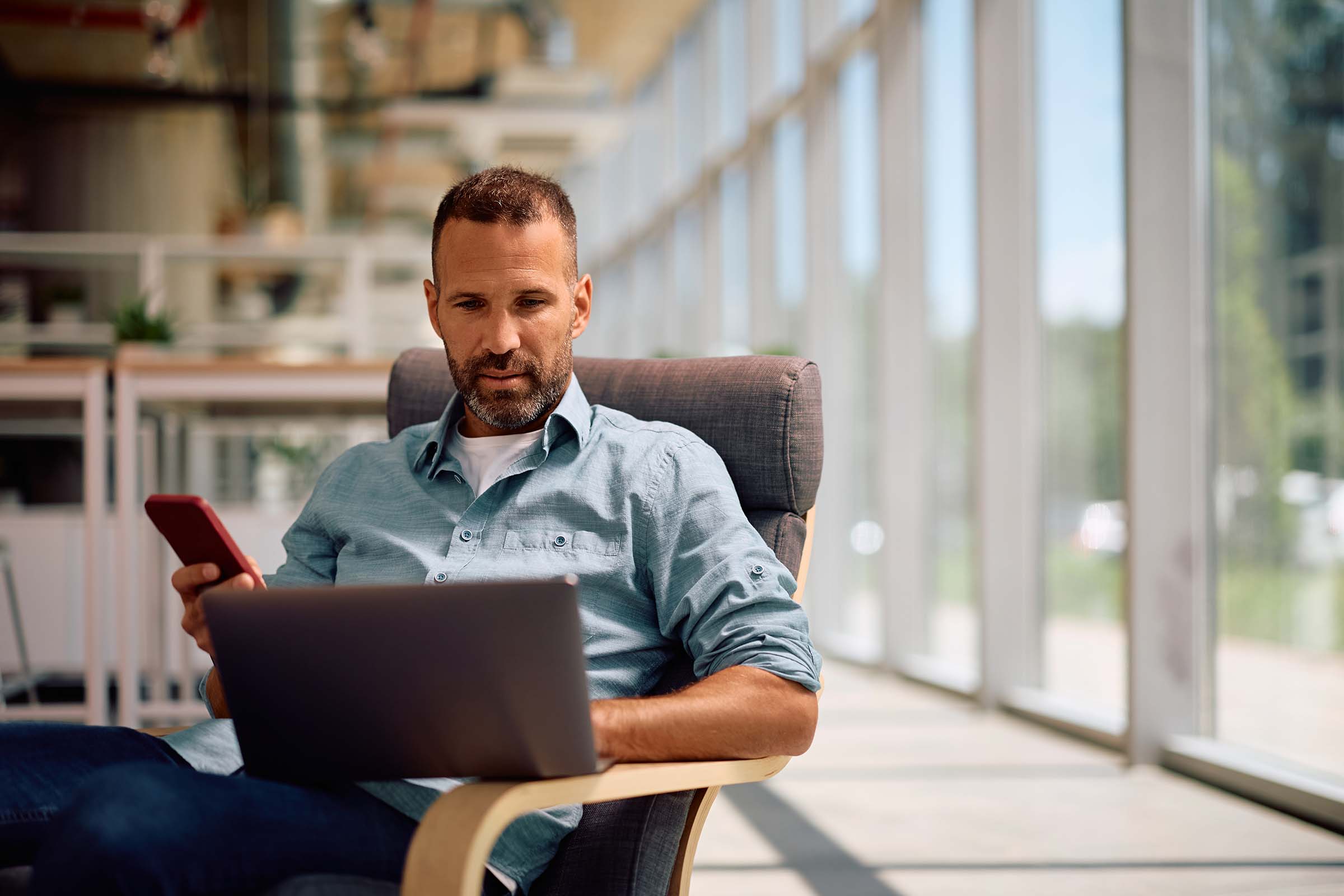 A man in a casual shirt sits in a bright, modern office space, holding a smartphone and working on a laptop.