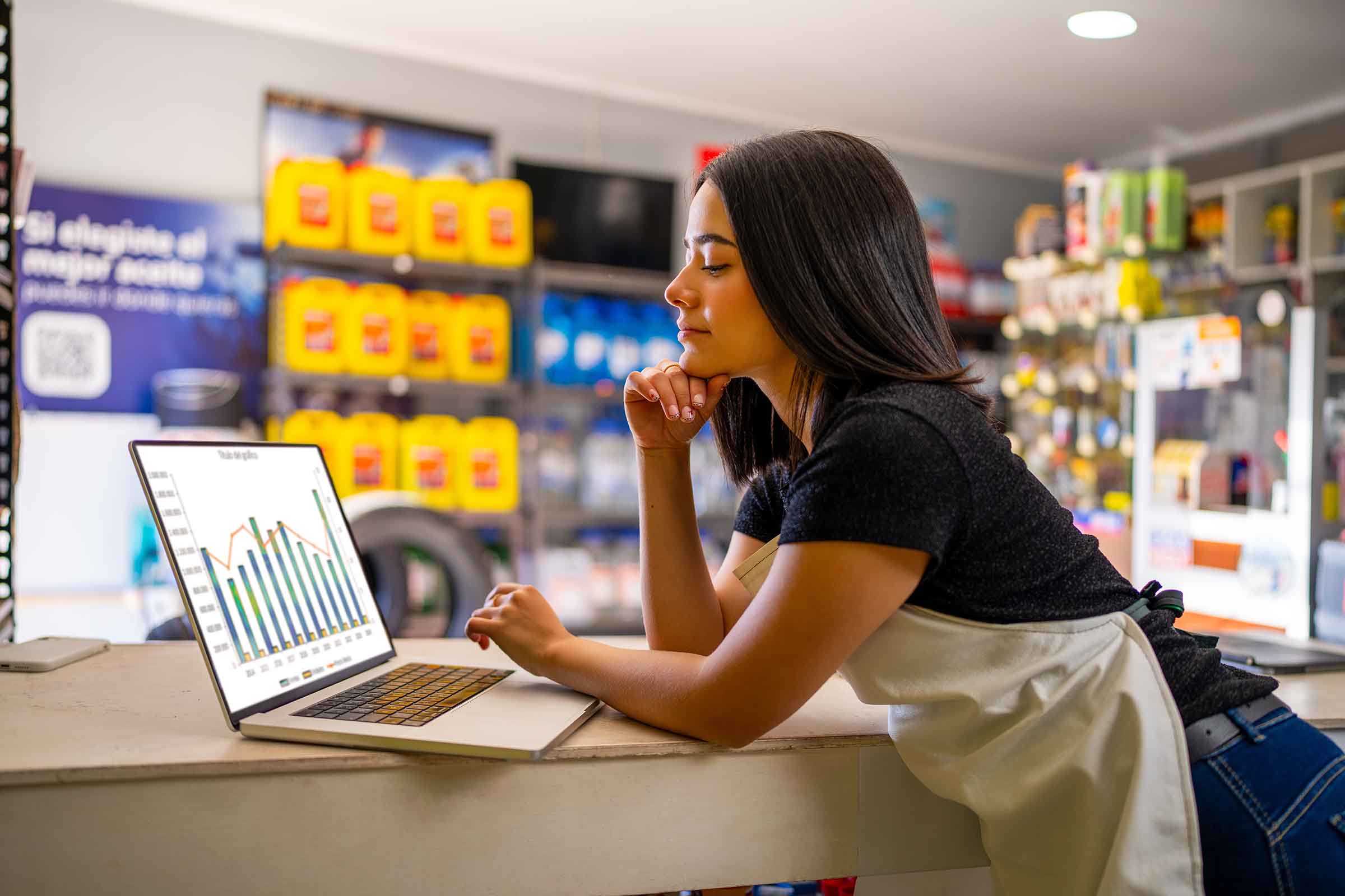 Woman in an apron at a store counter examining graph data on a laptop, with shelves of yellow bottles in the background.