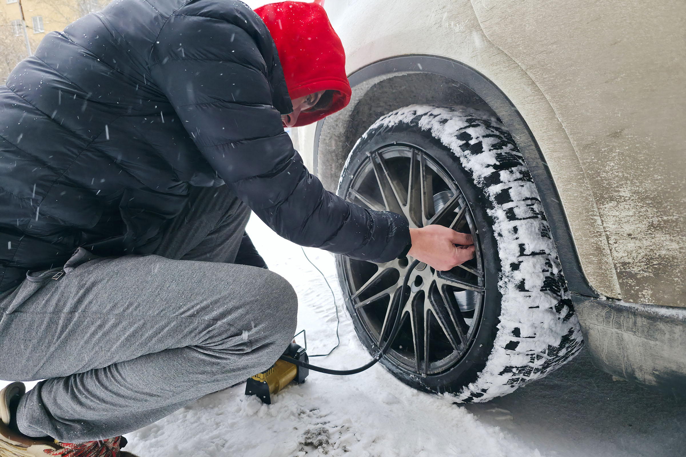 Person wearing winter clothing inflating a snow-covered car tire with a portable air compressor on a snowy day.