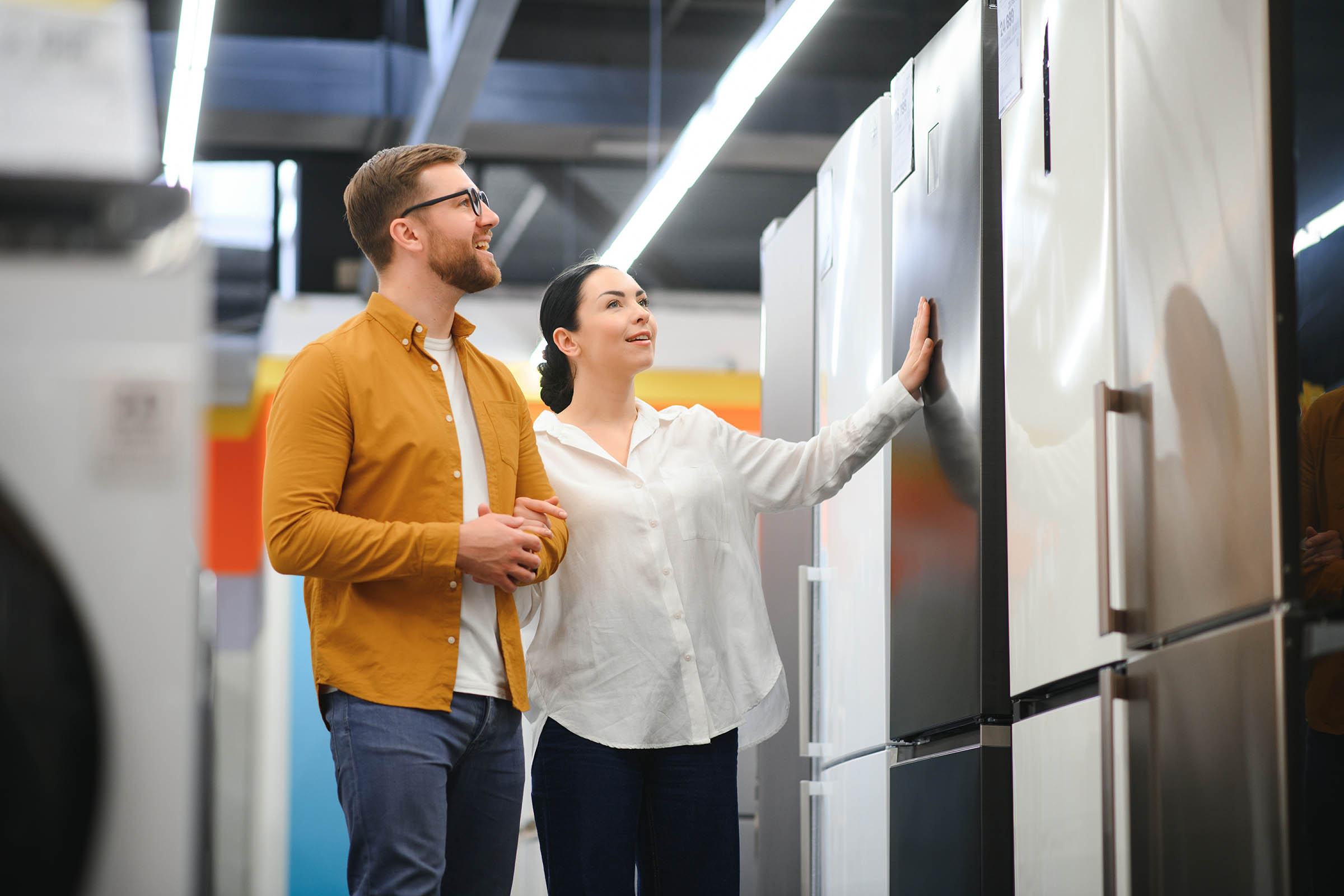 A couple examines refrigerators in a brightly lit appliance store, with colorful shelves in the background.