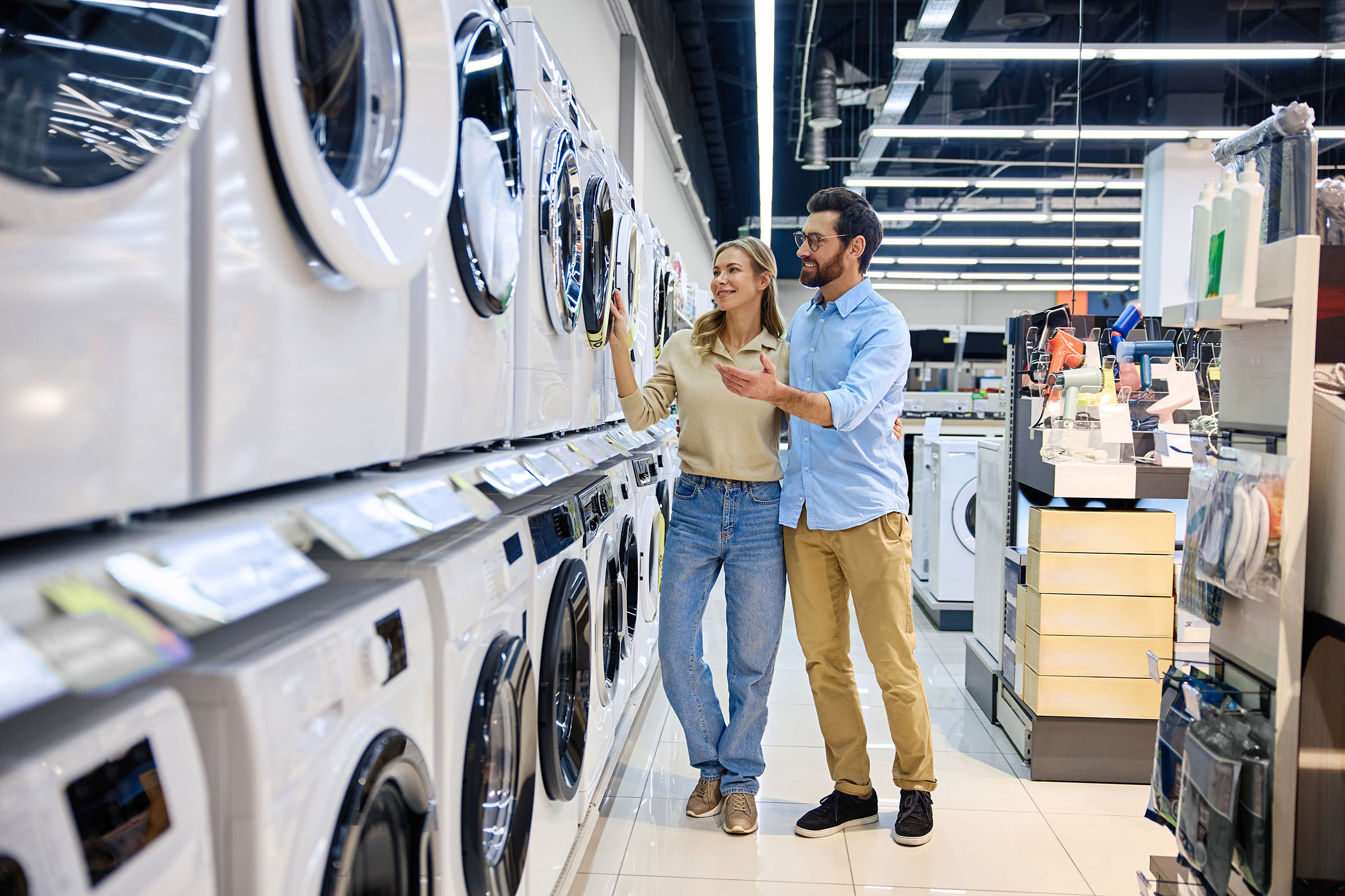 Smiling couple shopping for washing machines in an appliance store, examining front-loading models displayed in a row.