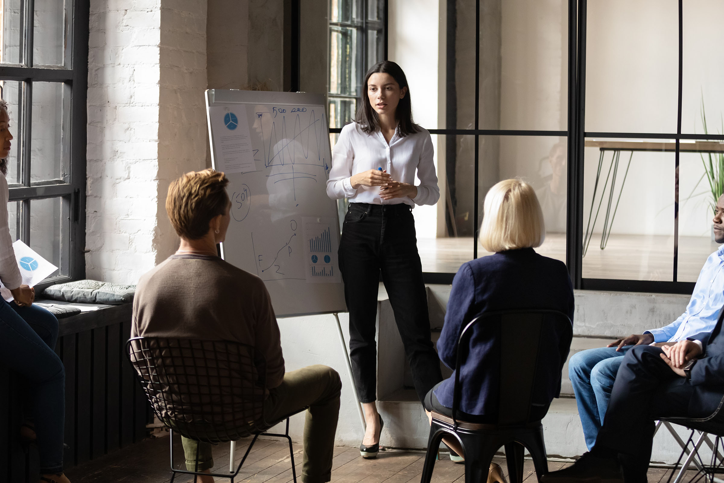 A woman stands by a whiteboard, presenting to a seated group in a modern office setting.