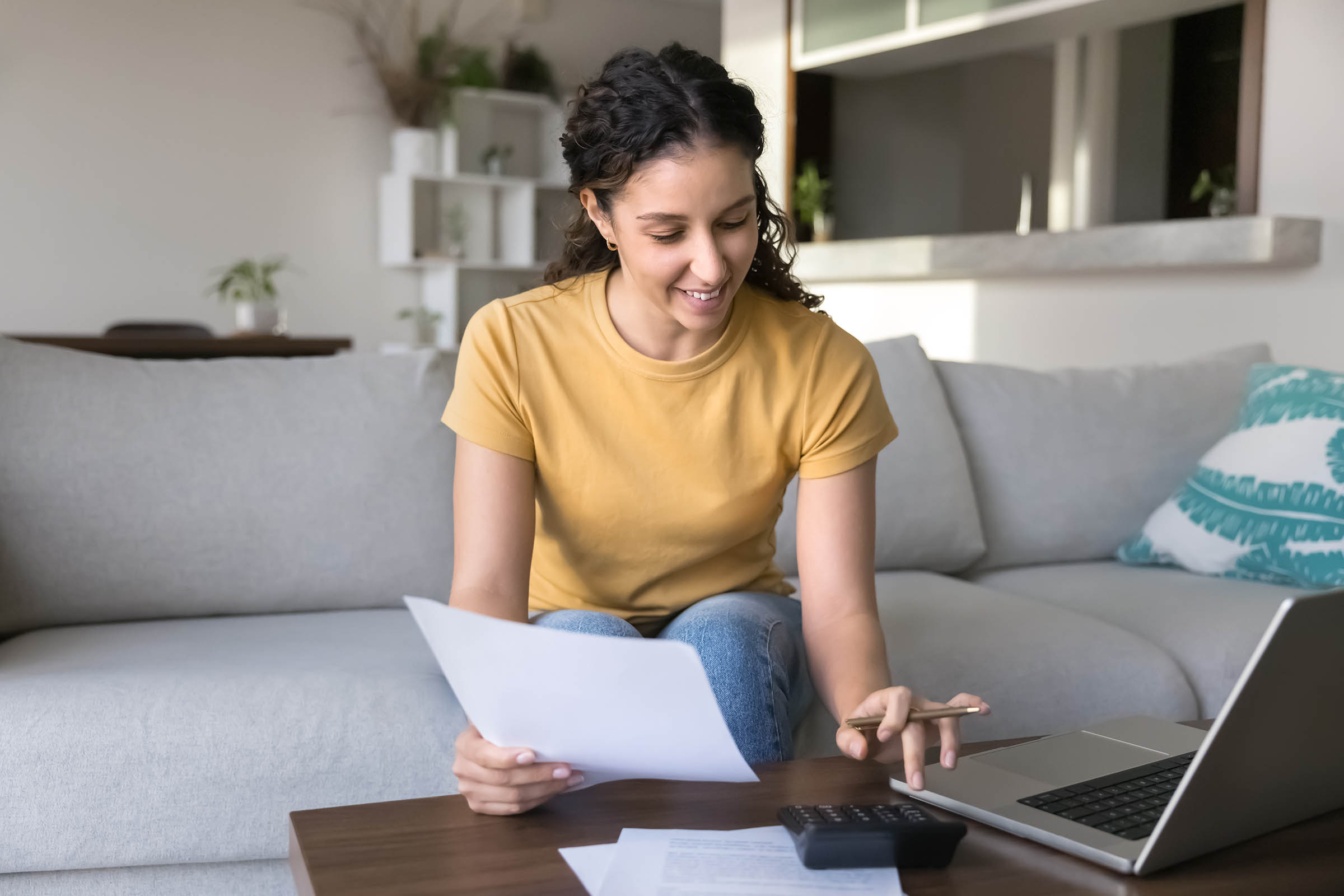 A woman in a yellow shirt sits on a couch, smiling as she reviews papers with a laptop and calculator on a table in front of her.