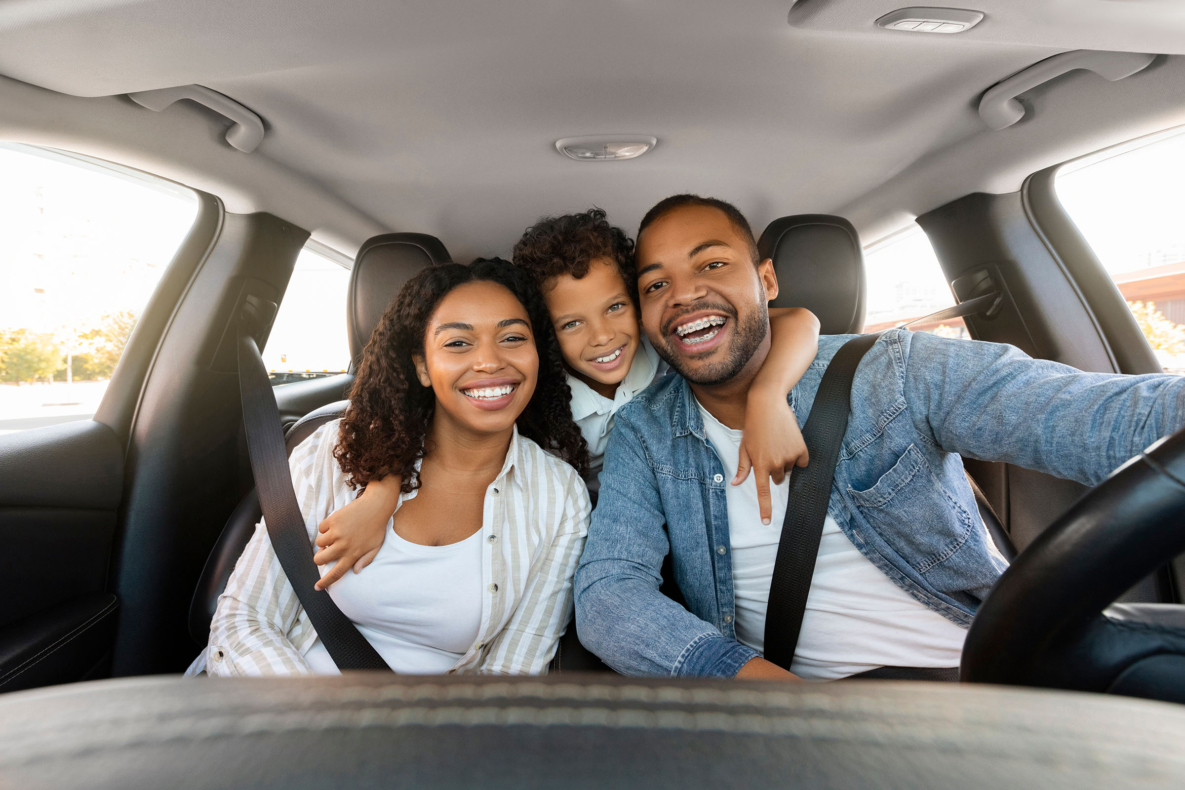 Smiling family of three sitting in a car, with a child leaning between two adults in the front seats, all looking at the camera.