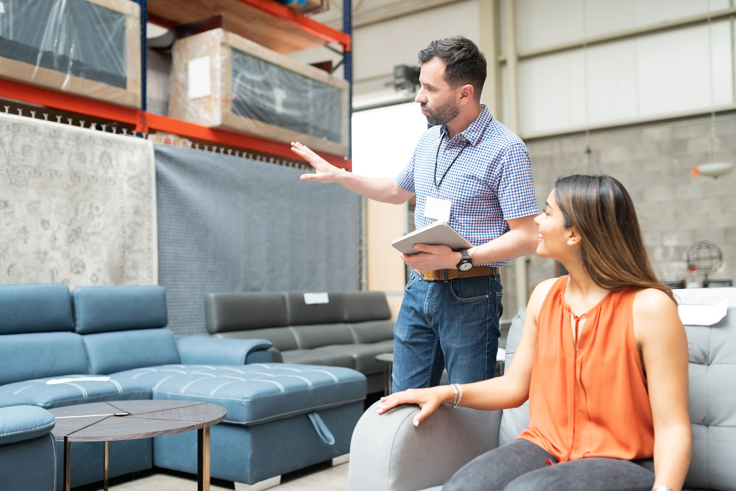 Furniture store employee holding a tablet and explaining sofa options to a seated customer.