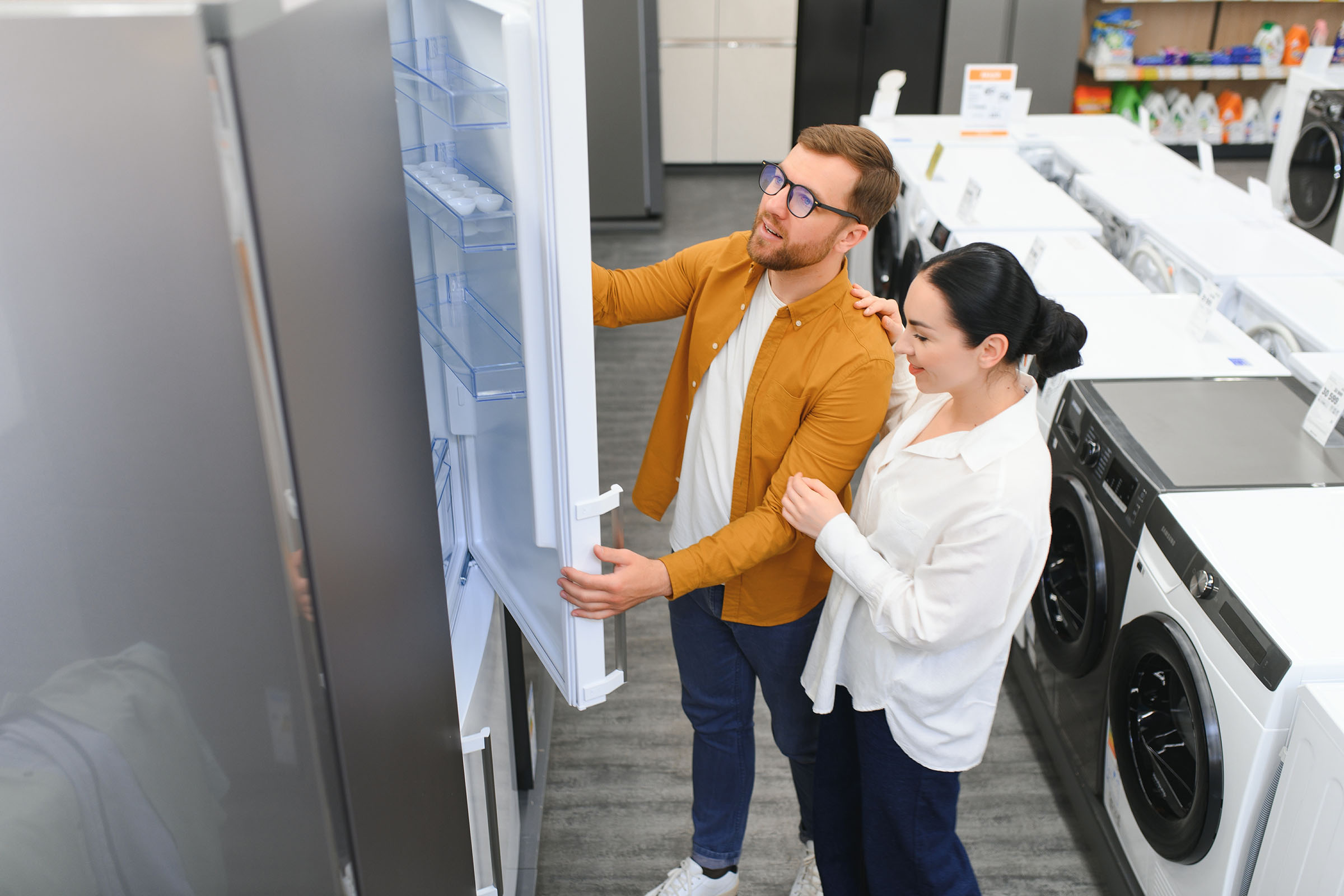 Couple inspecting the inside of an open refrigerator in an appliance store, standing near washers and dryers.