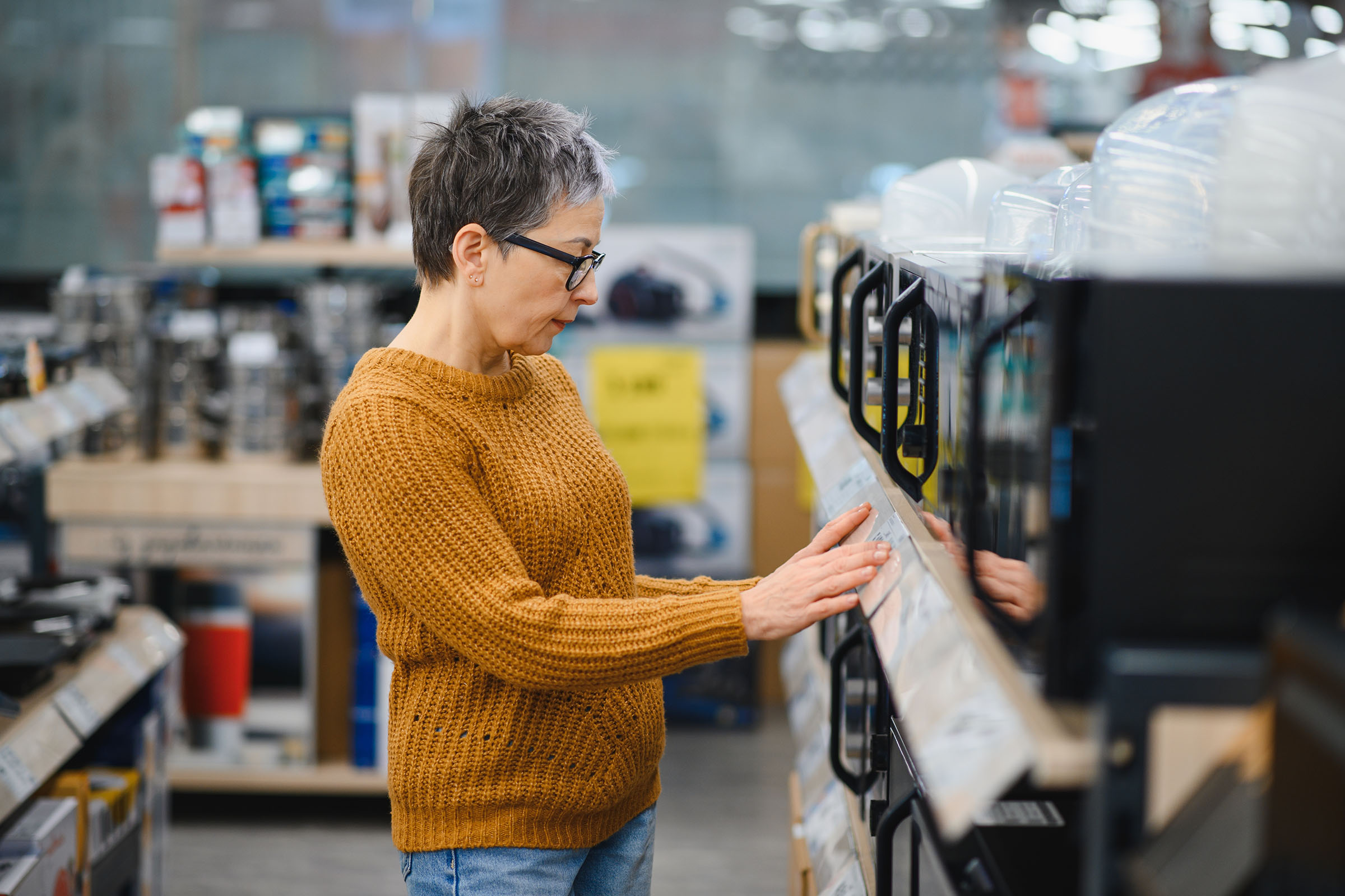 A woman in a mustard sweater examines appliances on display in a store.