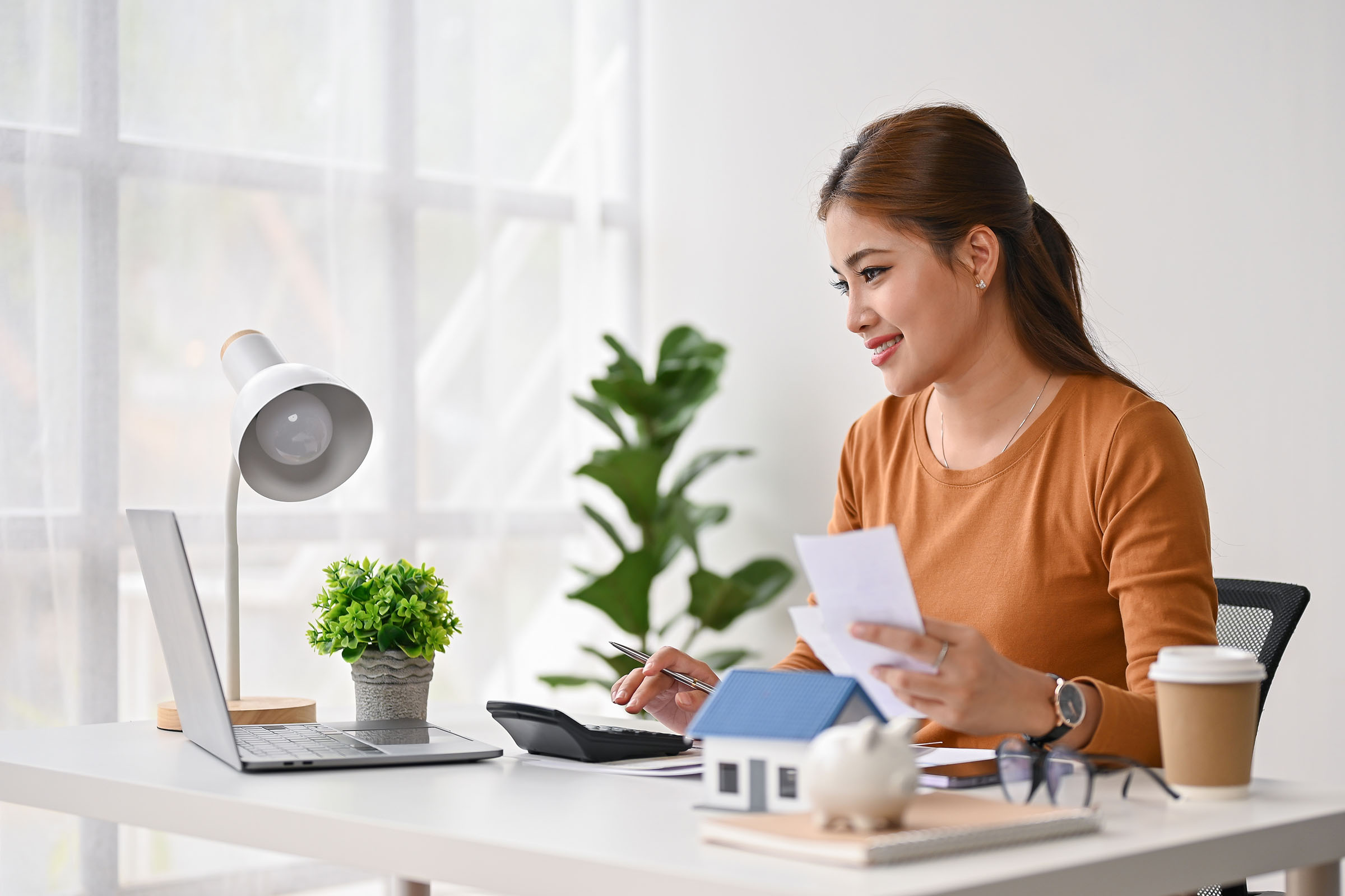 Smiling woman sitting at a desk using a calculator and holding papers, with a laptop, coffee cup, and small house model nearby.