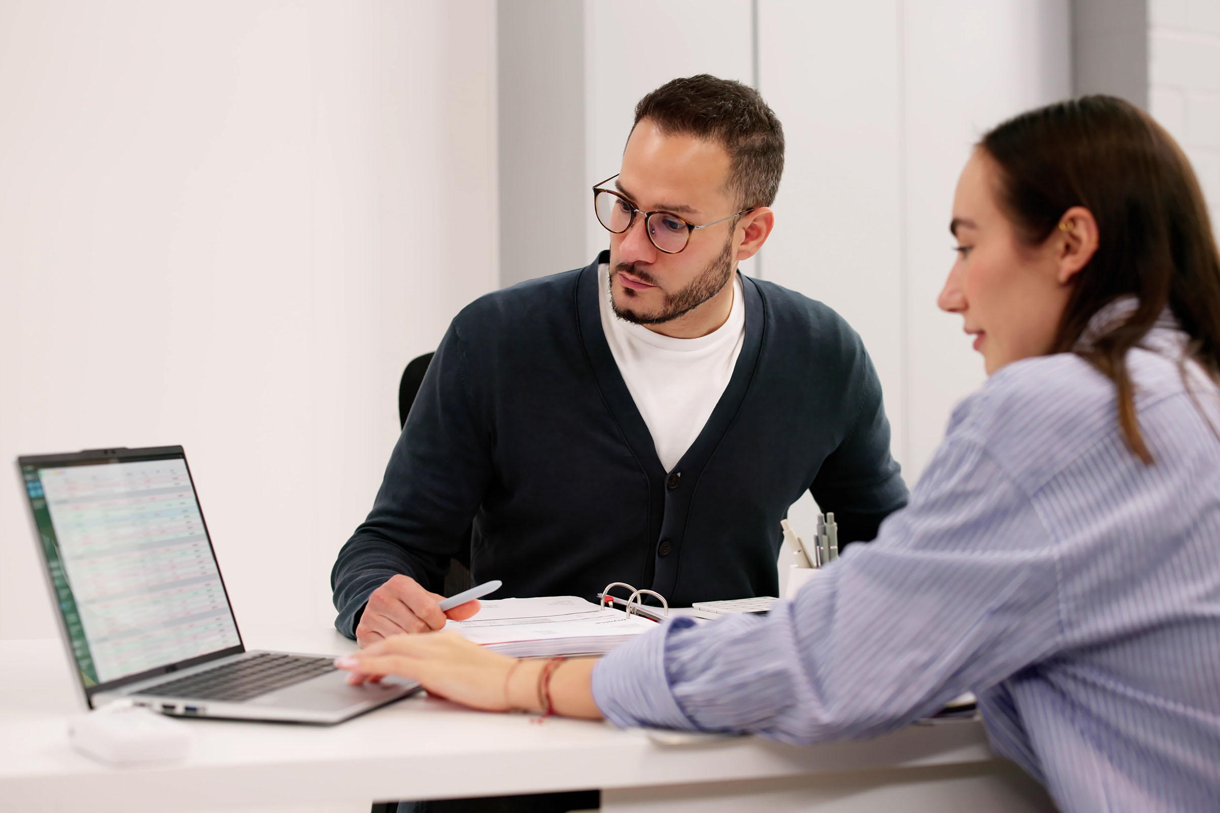 Two people discussing at a desk, one pointing at a laptop screen displaying spreadsheets, with papers and a pen nearby.
