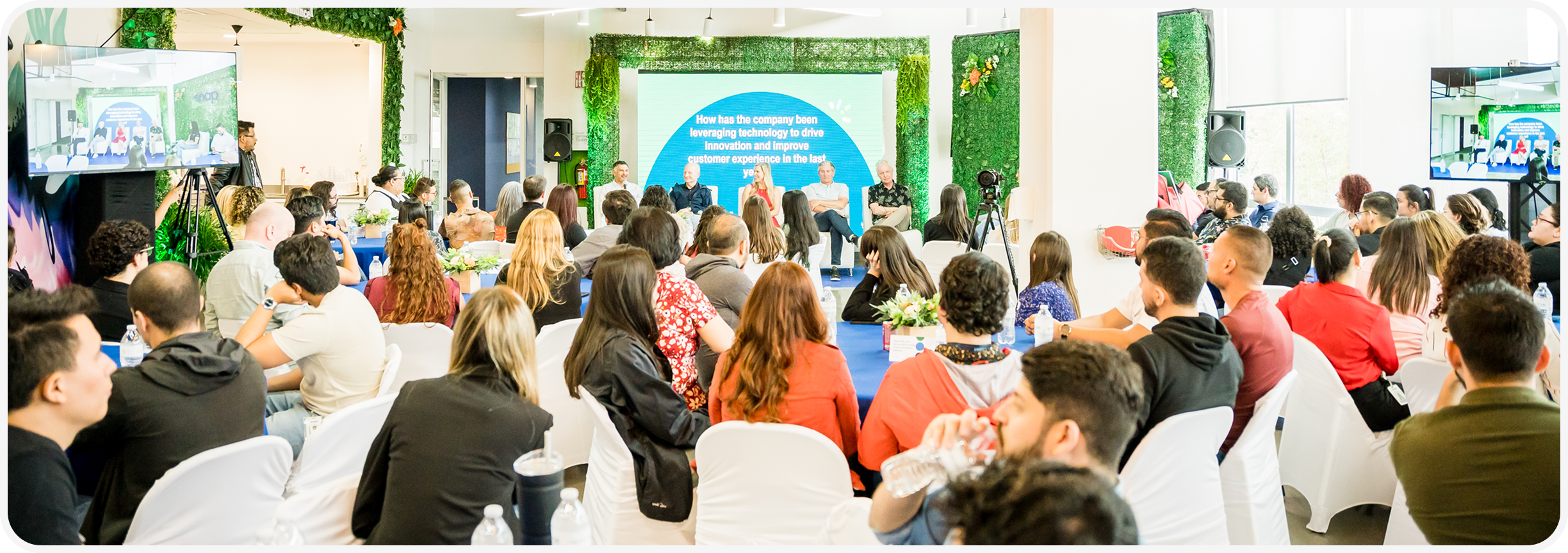 A diverse audience seated in a brightly lit room listens to a panel discussion. Screens display presentations, and greenery decorates the space.