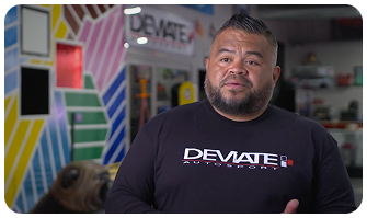 Man in black "DEVIATE Autostyling" shirt stands in a colorful, striped room with automotive decor.