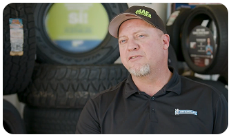 A man in a cap and polo shirt stands in front of stacked tires, with a blurred tire display in the background.