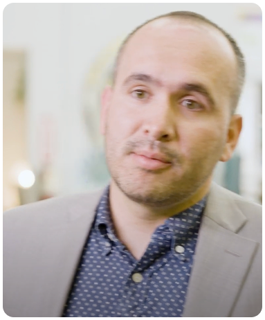 A person with short hair and a beard, wearing a gray blazer over a patterned shirt, standing indoors with a blurred background.