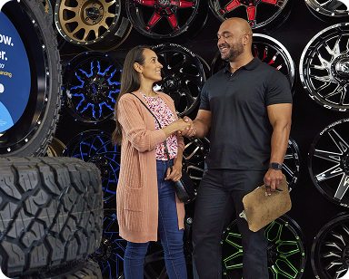 Two people smiling and shaking hands in front of a wall displaying various car rims, with large tires visible in the foreground.
