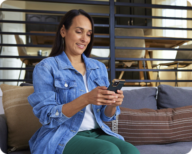 Woman in a denim jacket sits on a sofa, smiling as she looks at her smartphone. The room features a wooden dining table in the background.